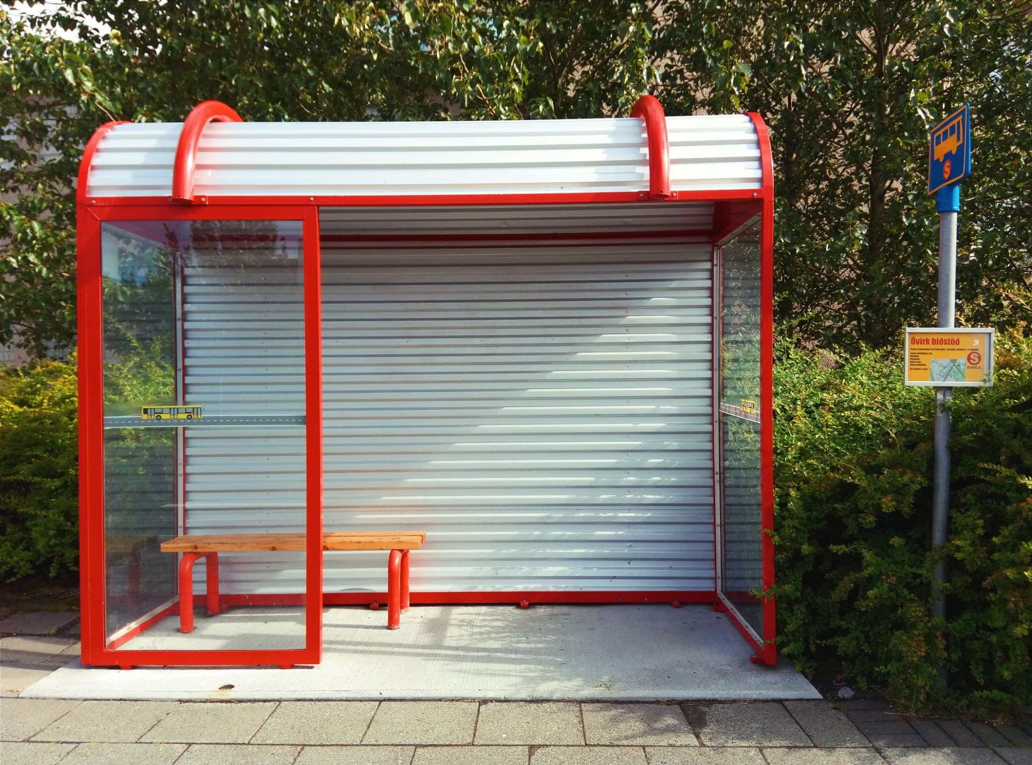 A red-framed bus stop shelter with a white corrugated roof and glass panels houses a wooden bench on a paved path next to a signpost with route information.