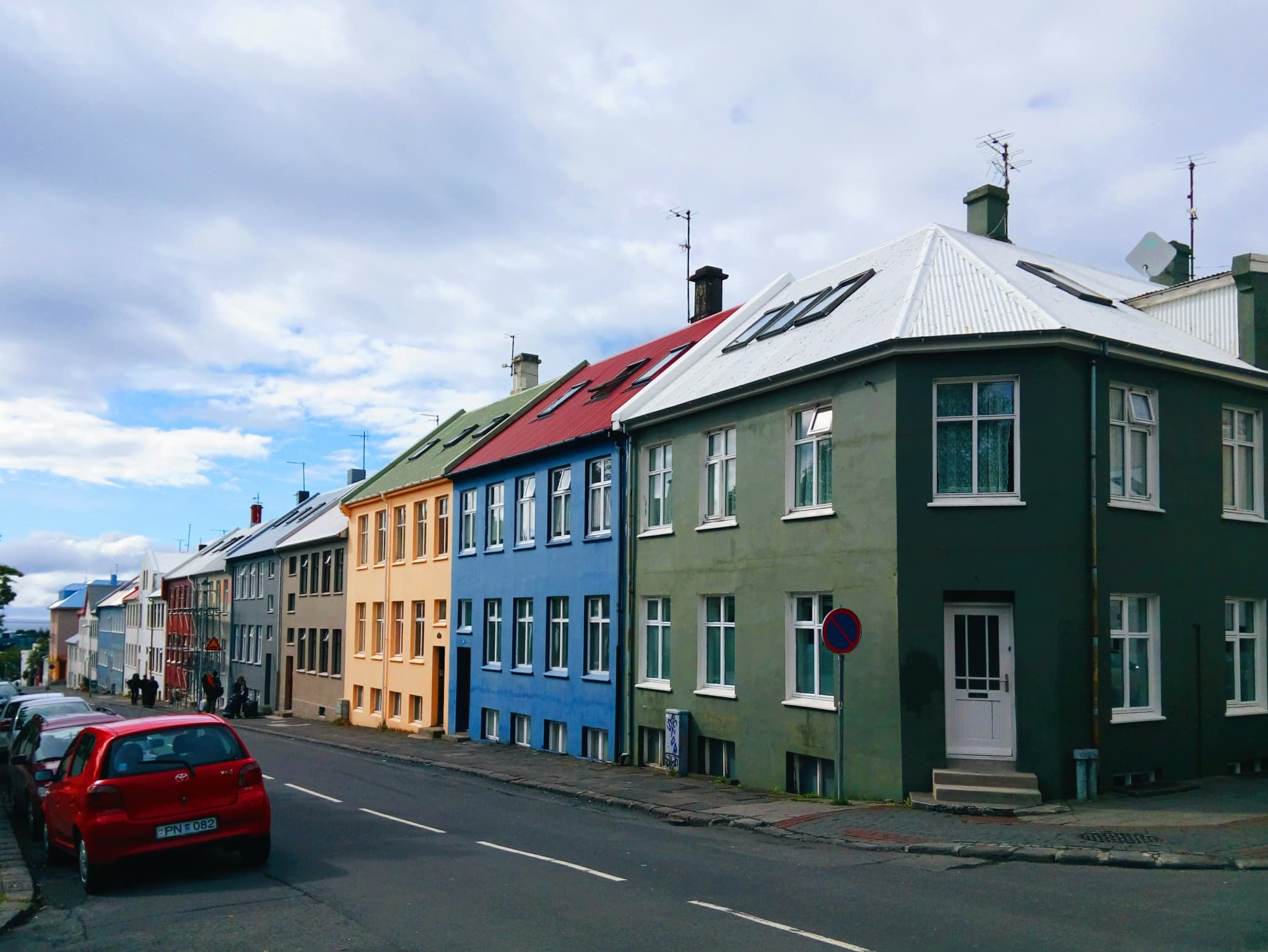 A row of colorful, multi-story buildings in shades of green, blue, yellow, and grey lines a paved street with a red car parked on the left side under a cloudy sky.