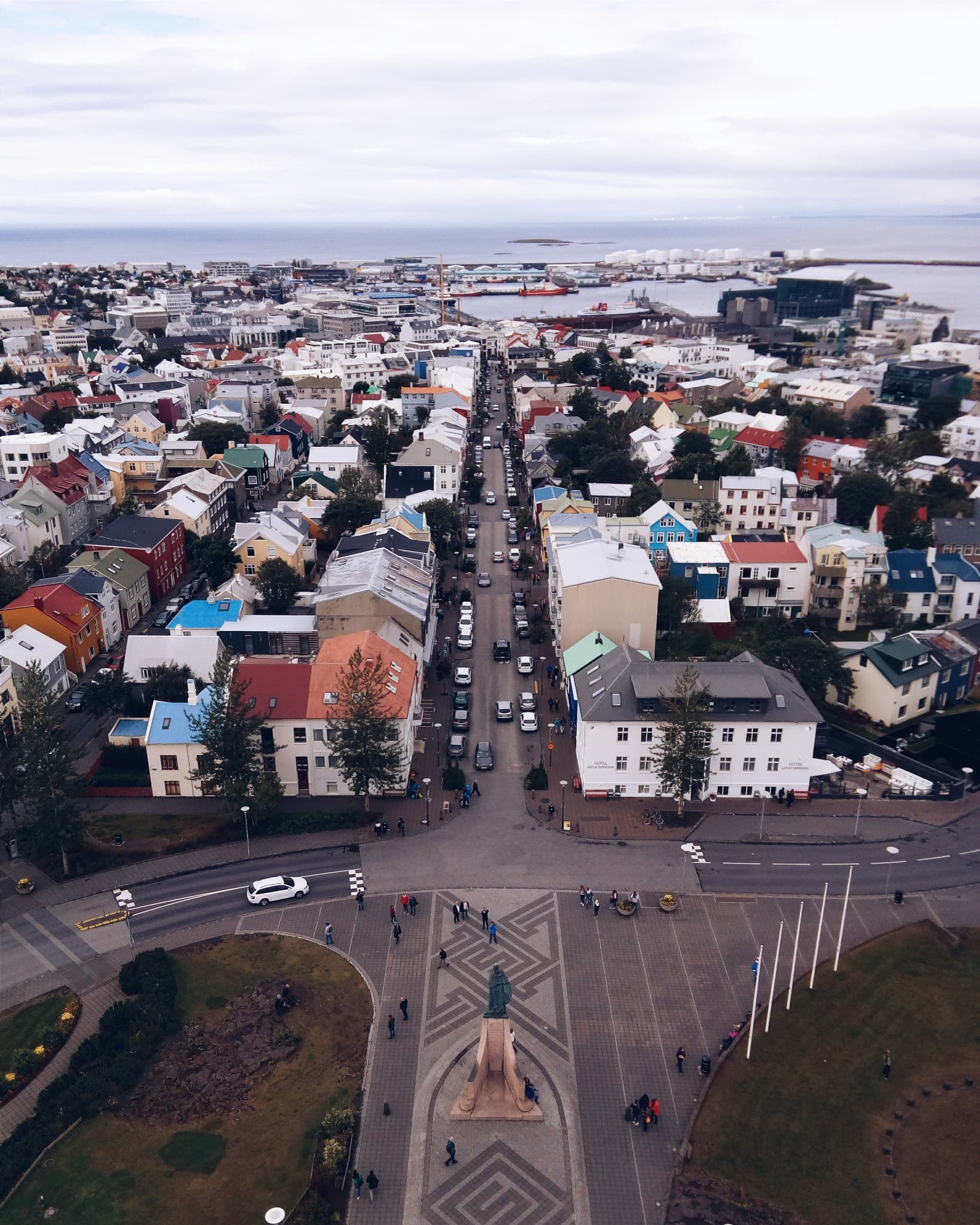An aerial view of a colorful city featuring a central plaza with a statue, streets lined with multi-colored houses, and a harbor in the background.