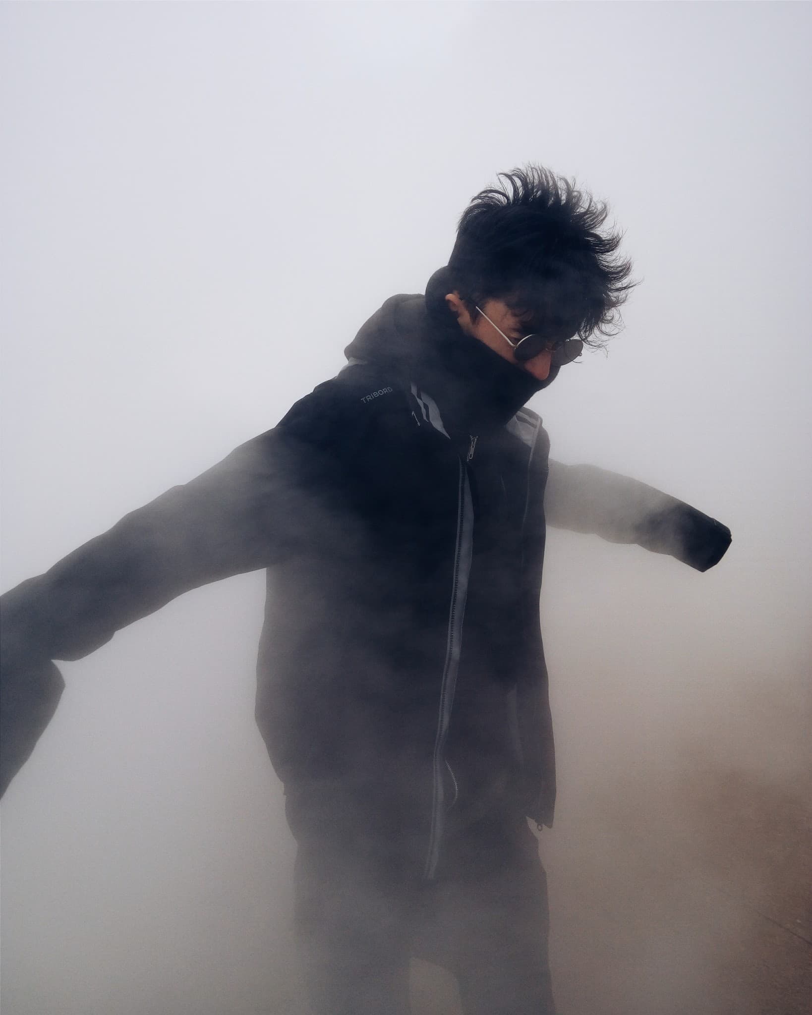 A person wearing a black jacket and sunglasses stands with arms outstretched amidst thick, white fog.