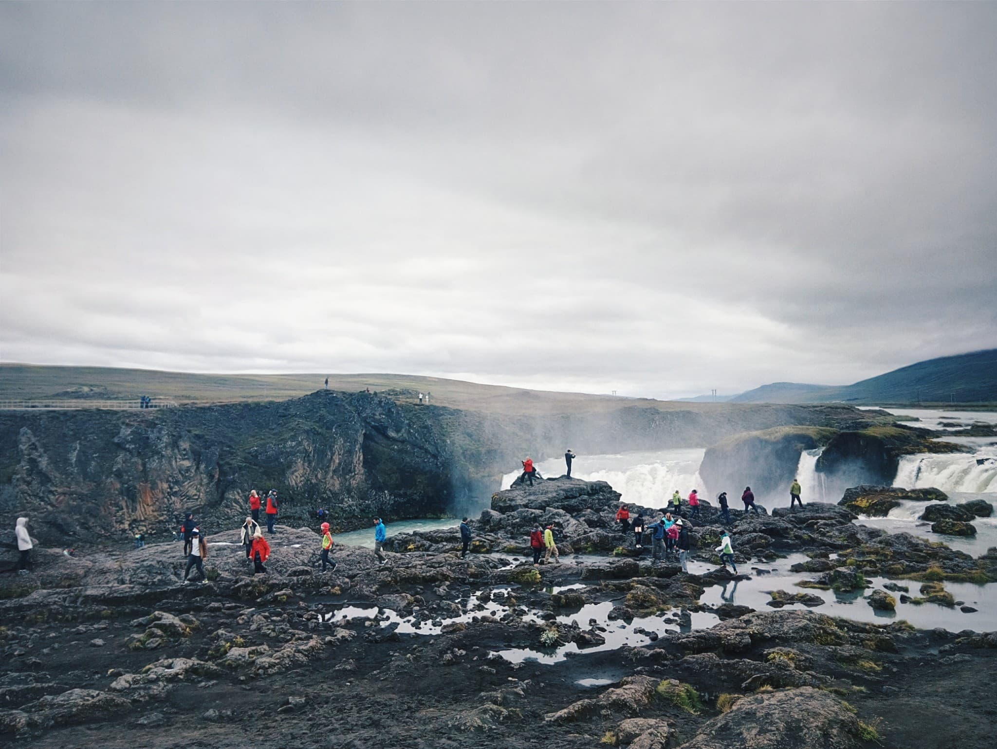 Tourists stand on rocky ledges at the base of a wide, powerful waterfall under an overcast sky.