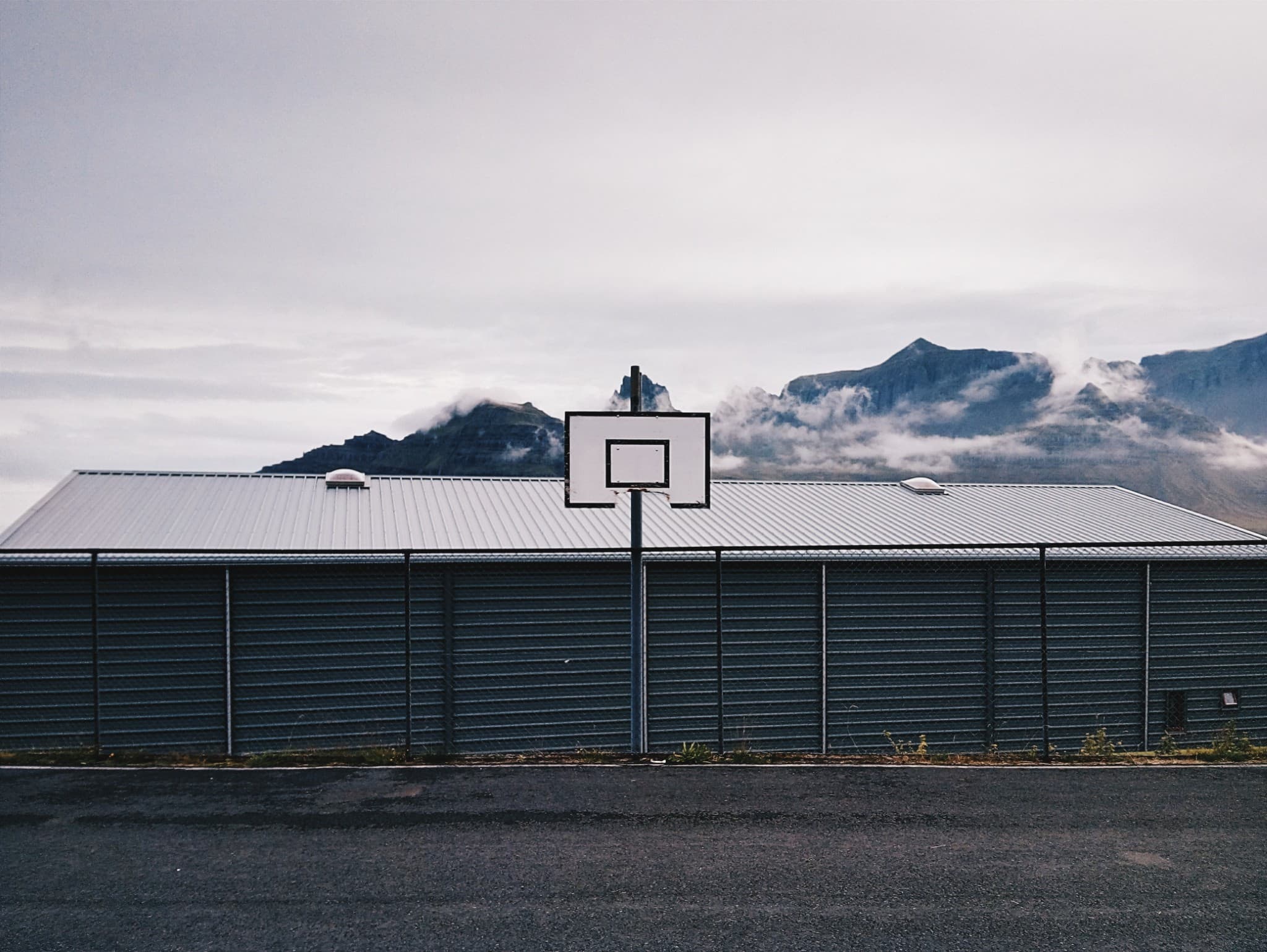 A basketball hoop stands in front of a long, dark metal fence, with misty mountains visible in the background under an overcast sky.