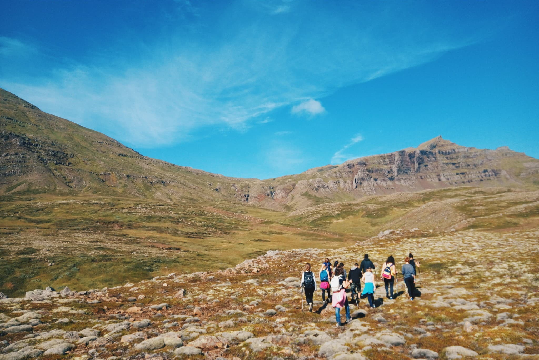 A group of hikers walks across a rocky, grassy landscape in a mountain valley under a bright blue sky.