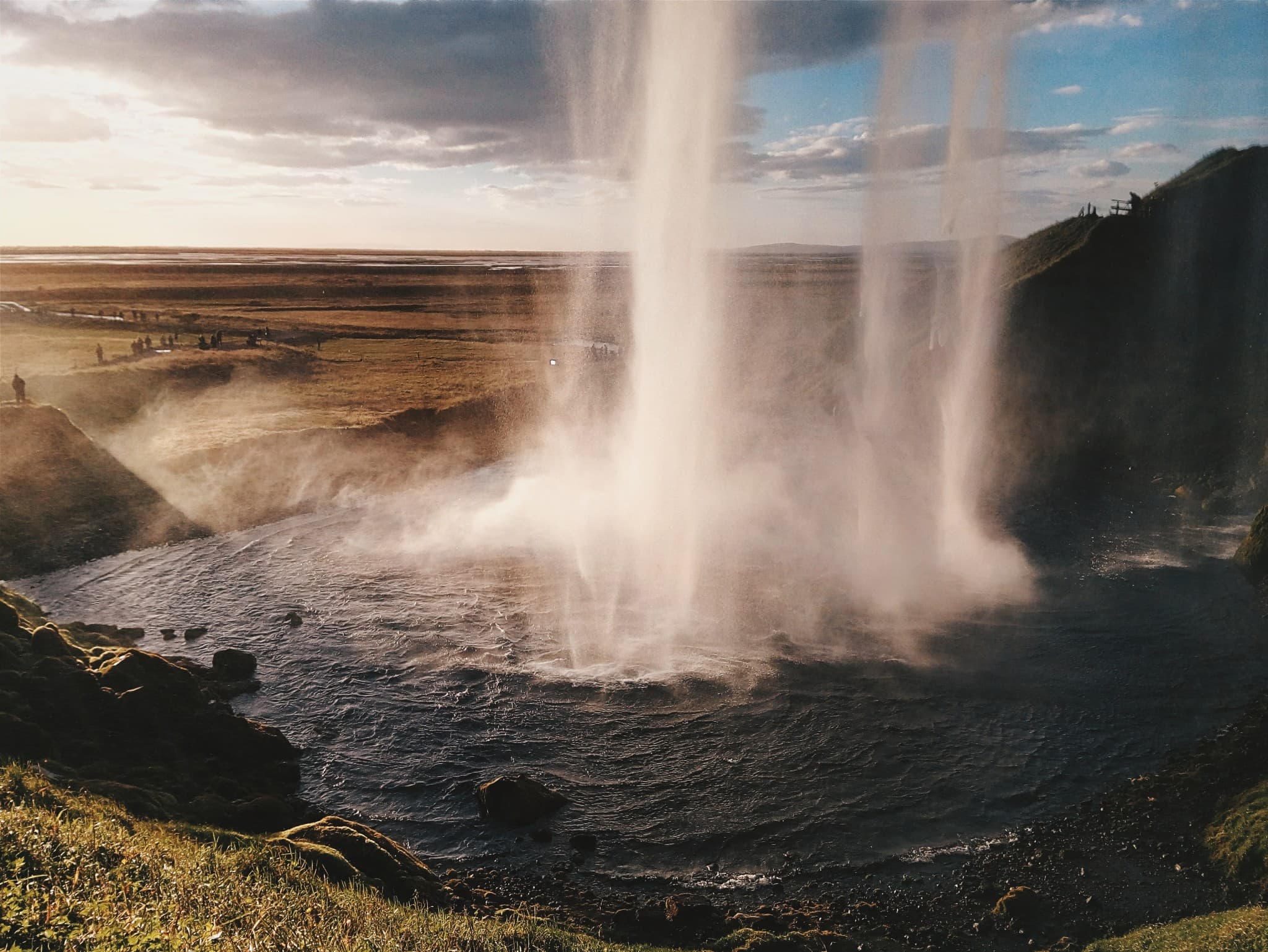 A wide waterfall crashes down into a river pool, sending mist into the air against a backdrop of flat fields and a partly cloudy sky.