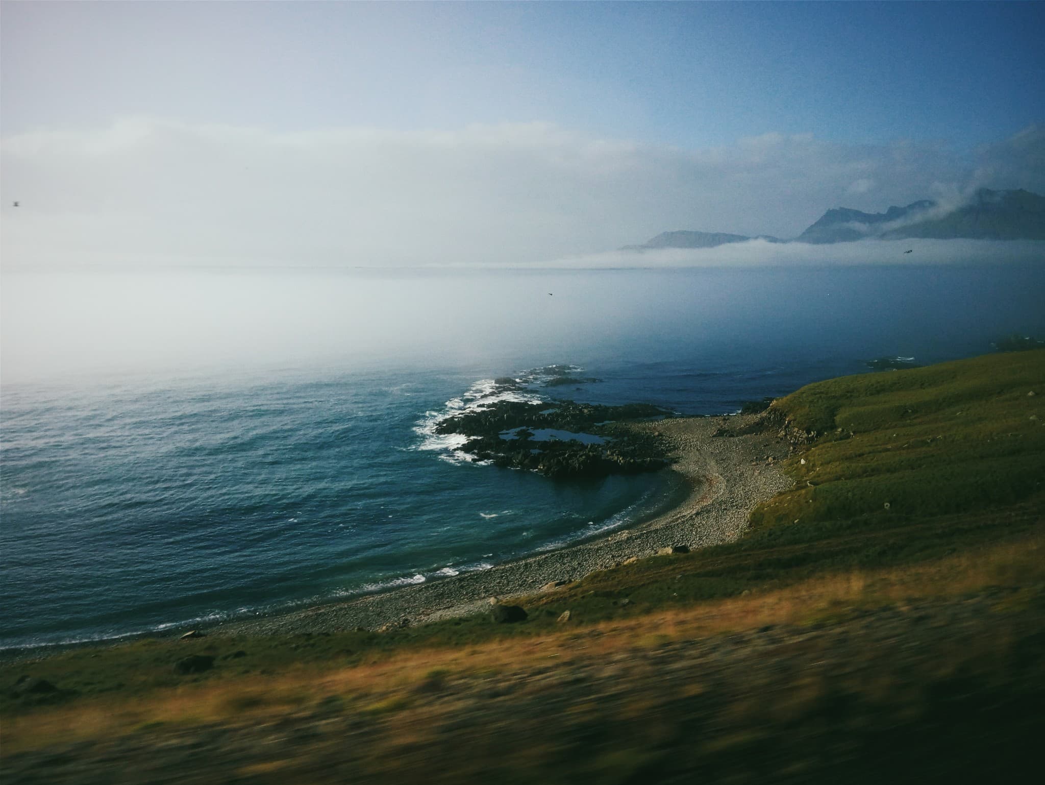A view of a rocky coastline and blue ocean with green hills in the foreground and distant mountains partially obscured by clouds.