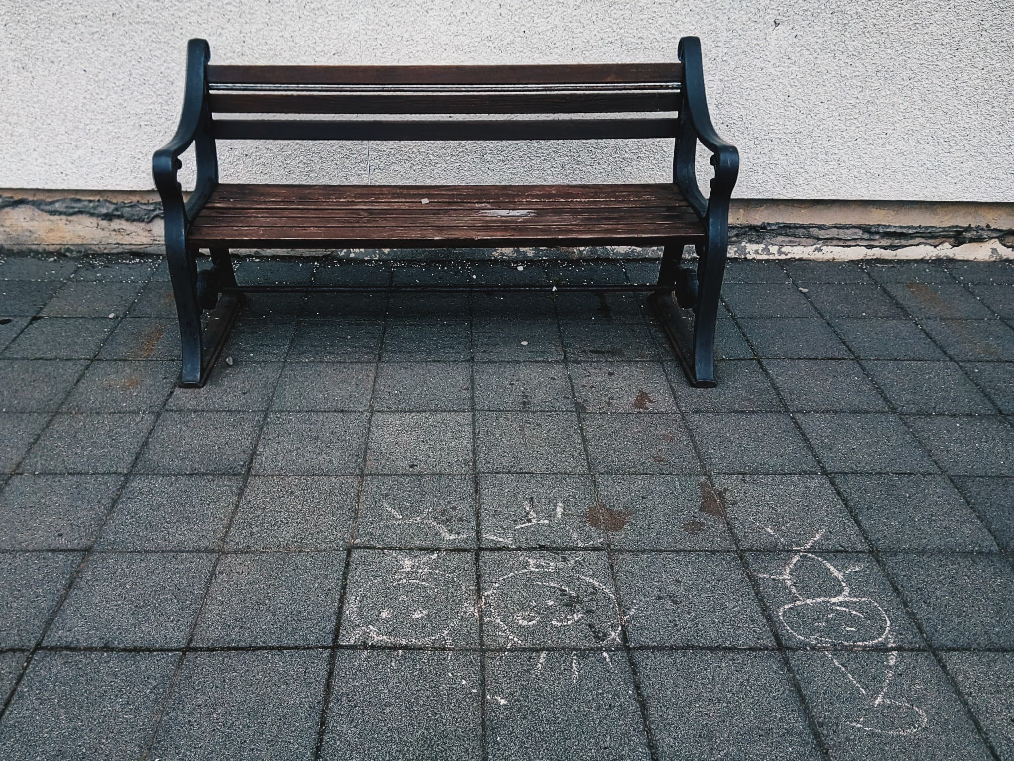 An empty wooden bench sits on a paved walkway against a white wall, with chalk drawings of a sun and a face drawn on the ground nearby.