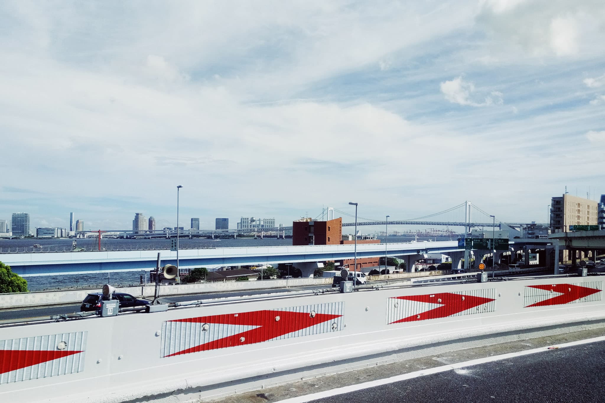 A view from a highway shows a concrete barrier with red and white arrows in the foreground, overlooking a bay with a large suspension bridge and city buildings in the distance.