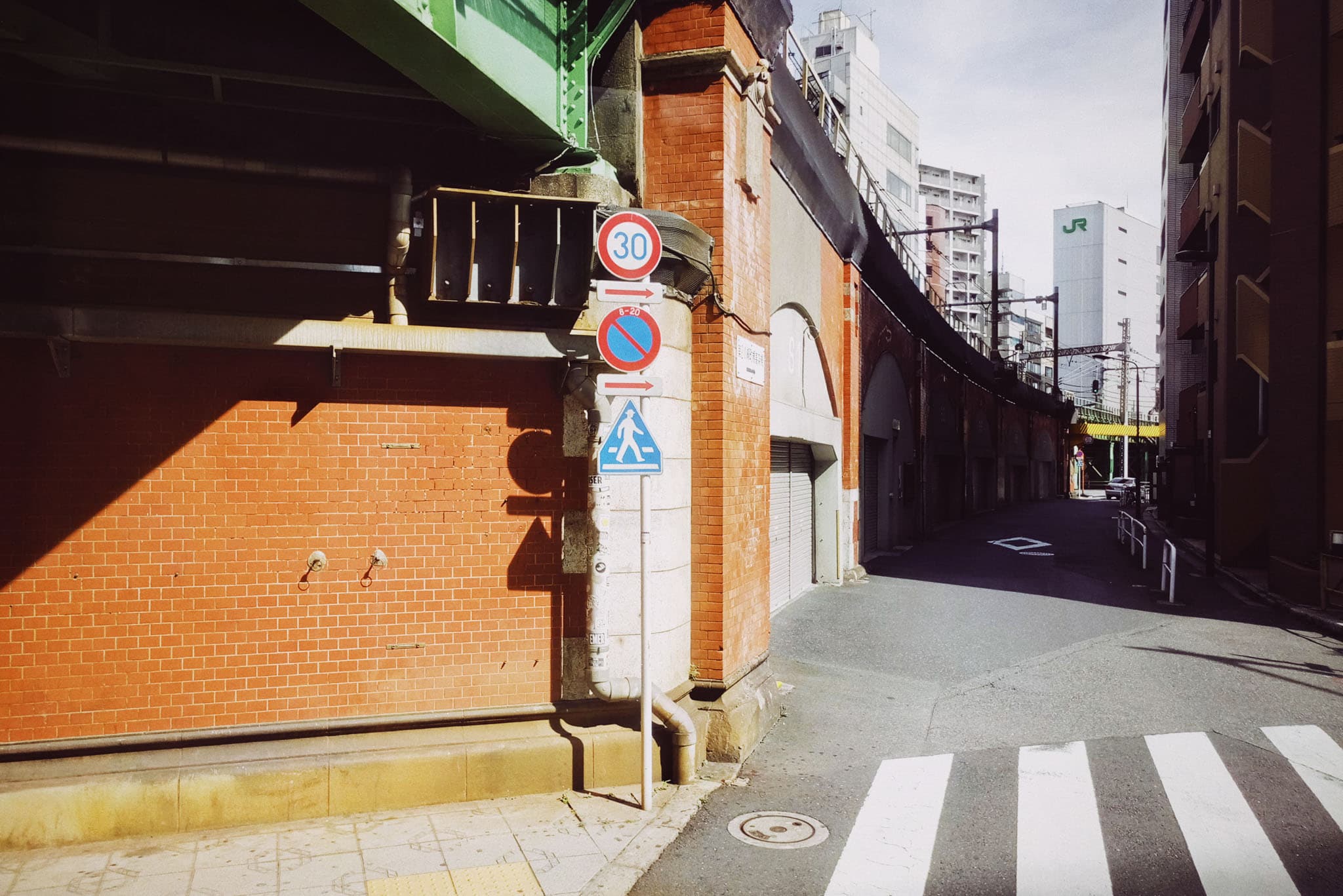 A sunlit street scene shows a red brick wall with traffic signs on the left, leading down a narrow road towards tall buildings in the distance.