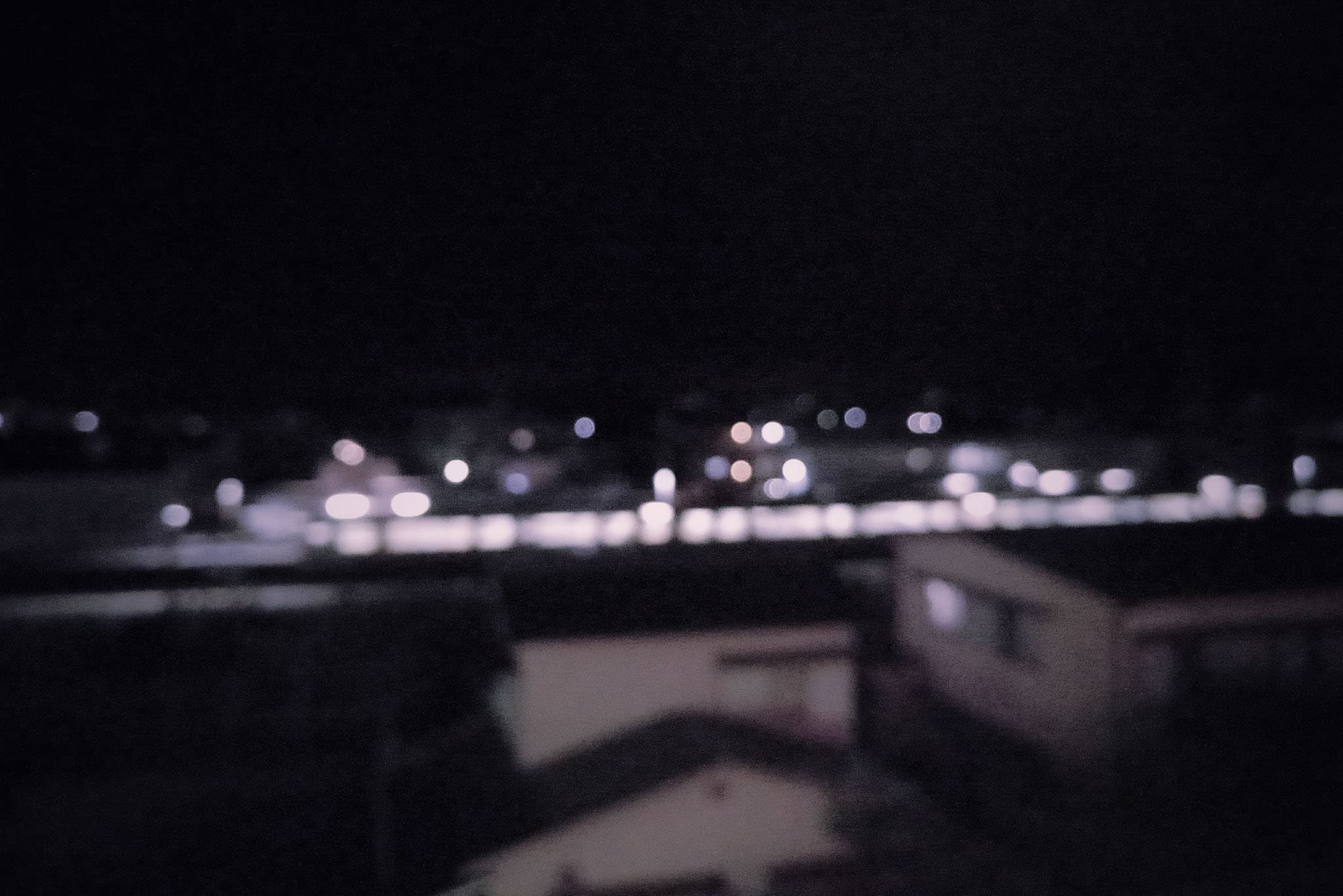 This is a blurry, out-of-focus night view overlooking a row of rooftops with distant, glowing lights.