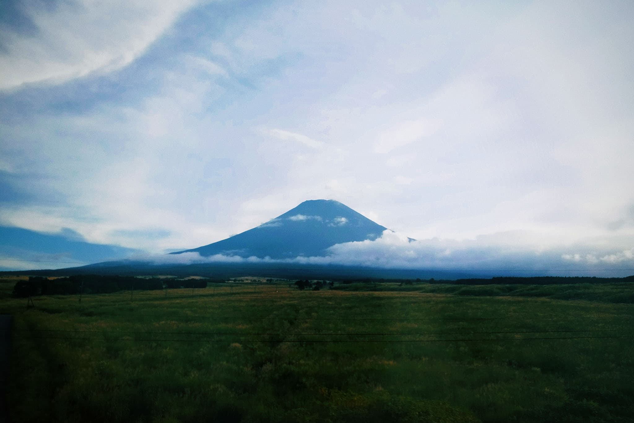 A large, cone-shaped mountain rises in the center of a wide, green landscape under a cloudy sky.