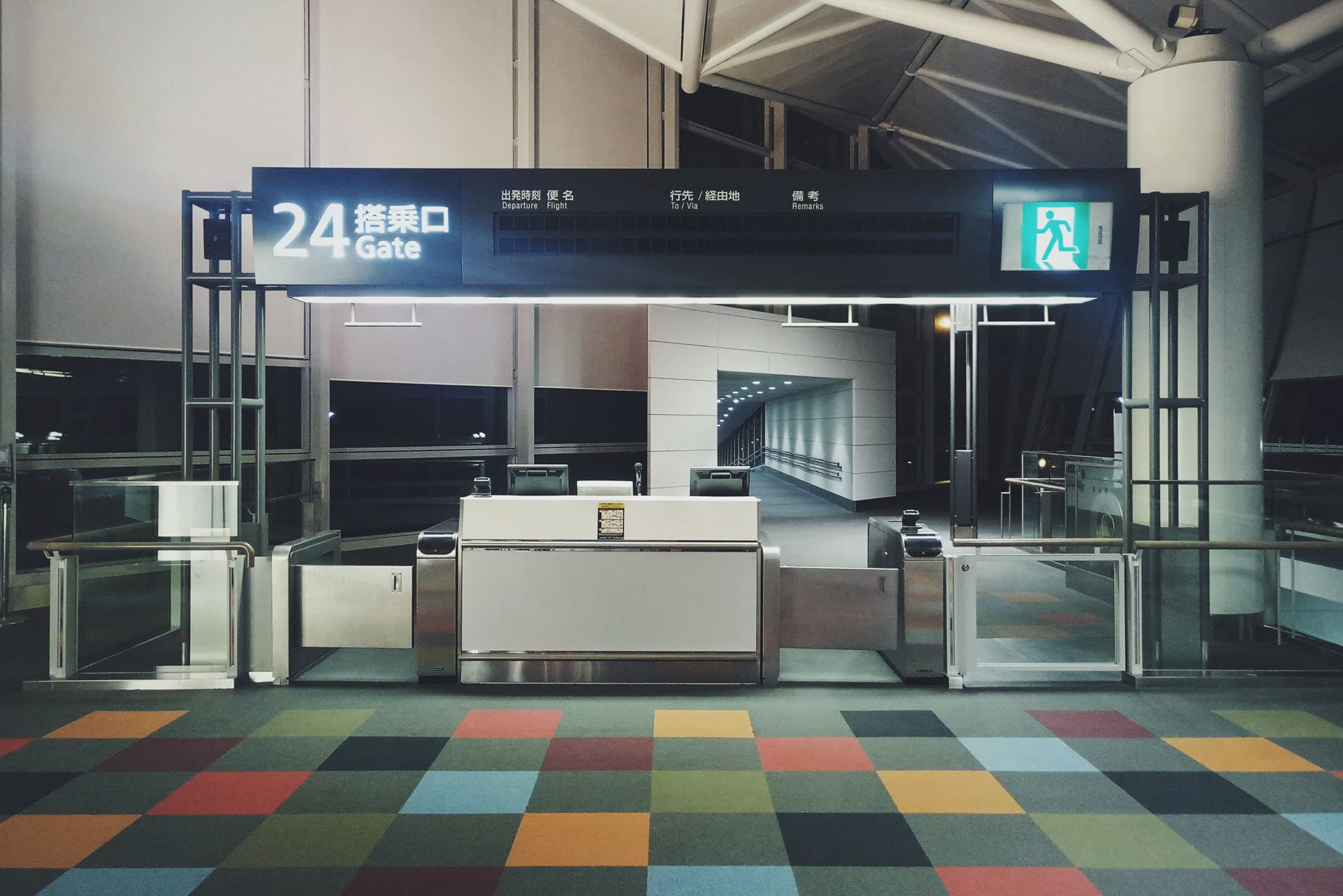 This image shows the entrance to gate 24 at an airport, featuring a large overhead sign, security turnstiles, and a colorful checkered floor.