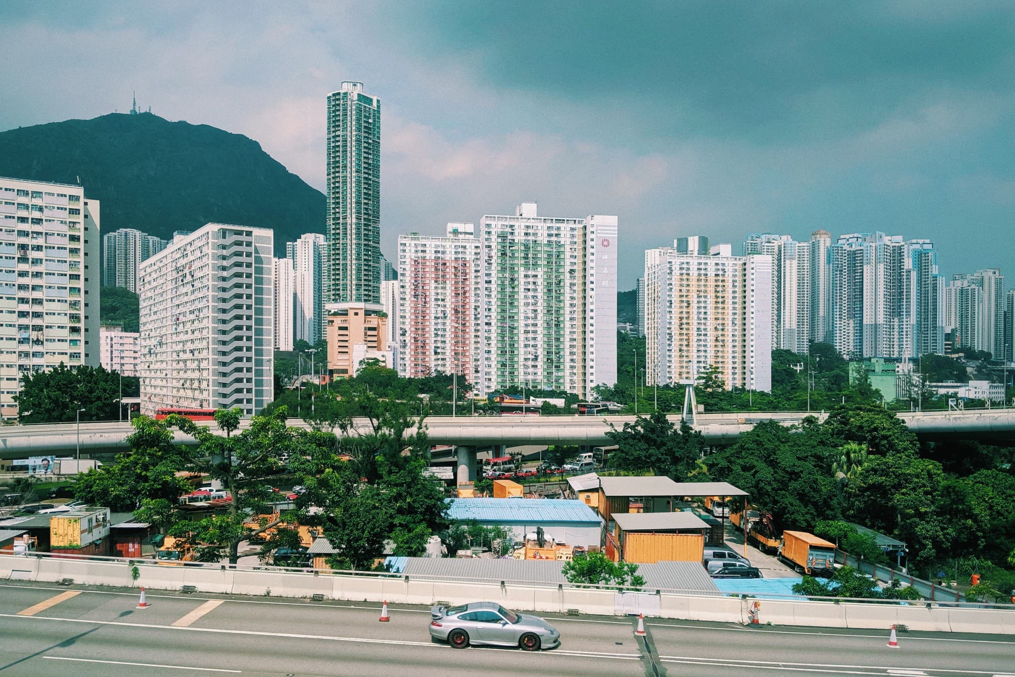 A silver car travels on a highway in the foreground, set against a backdrop of dense high-rise residential buildings and a large green mountain under a cloudy sky.