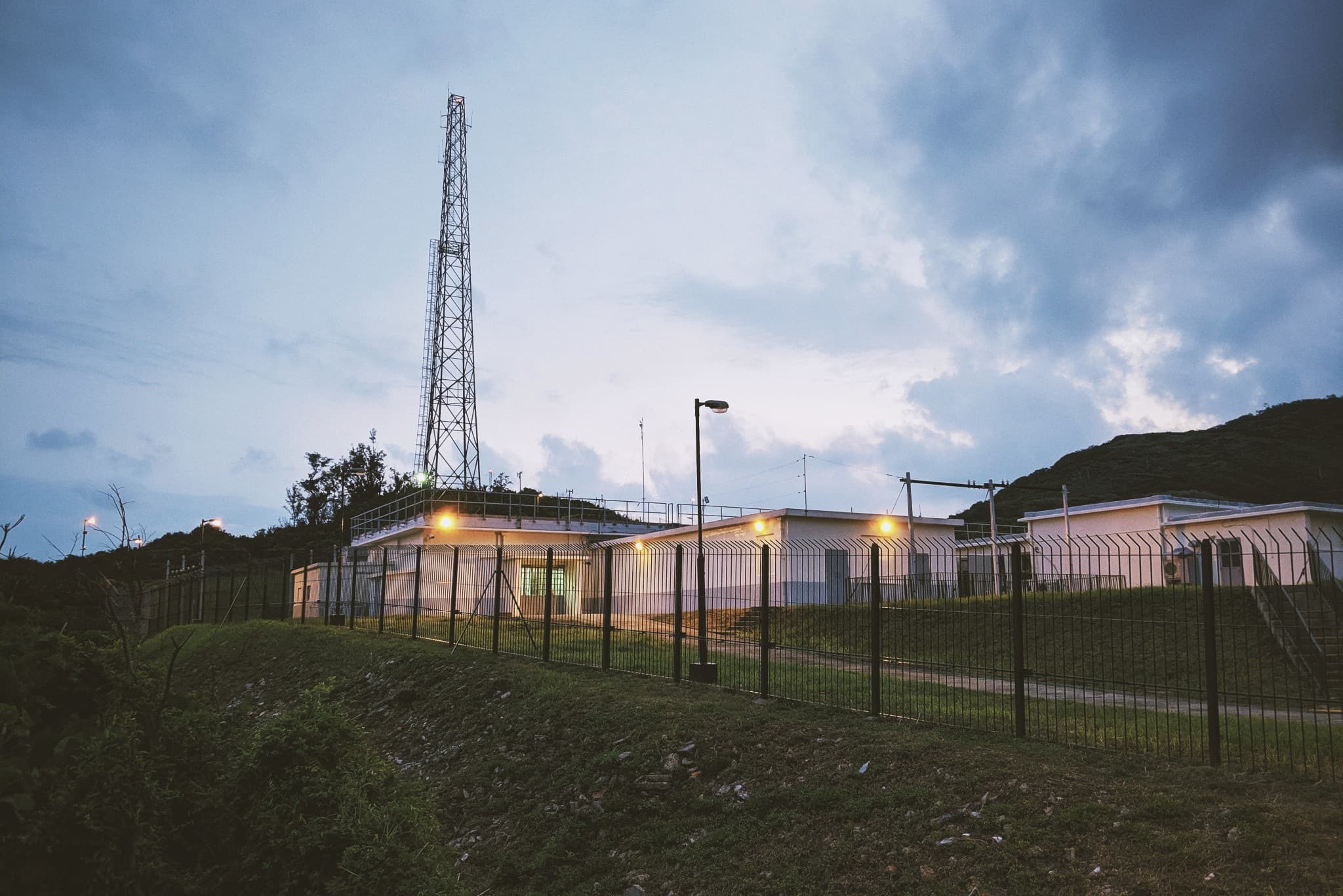 A tall metal lattice tower stands next to white industrial buildings behind a black fence, illuminated by warm lights against a cloudy twilight sky.