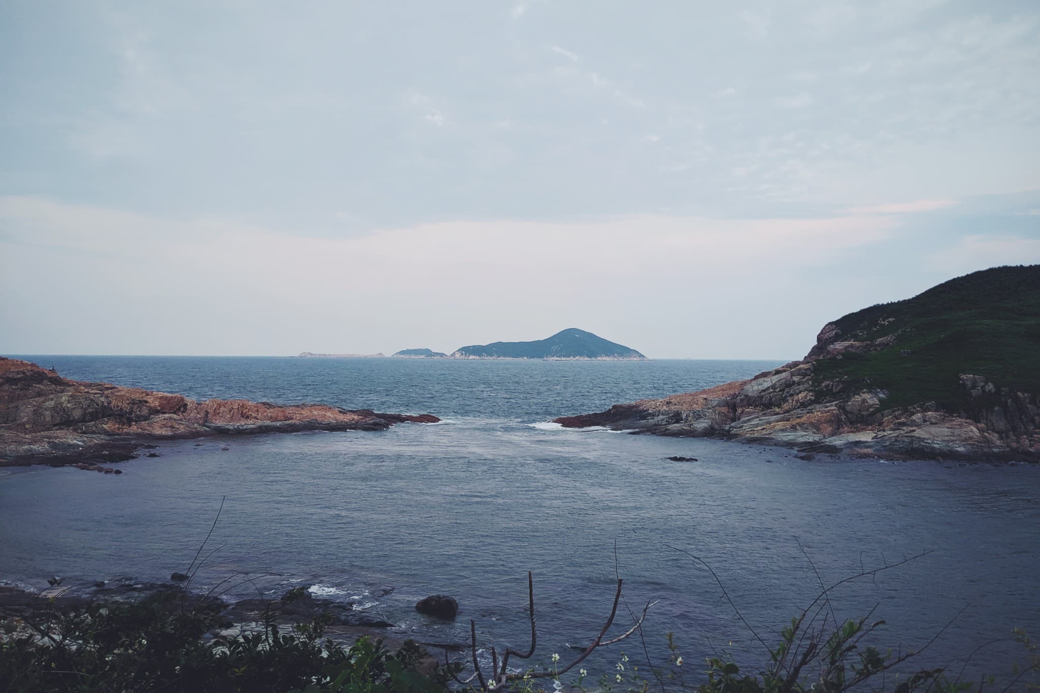 A rocky coastline frames a calm blue bay that opens up to the ocean with small, hazy islands visible on the horizon under a cloudy sky.