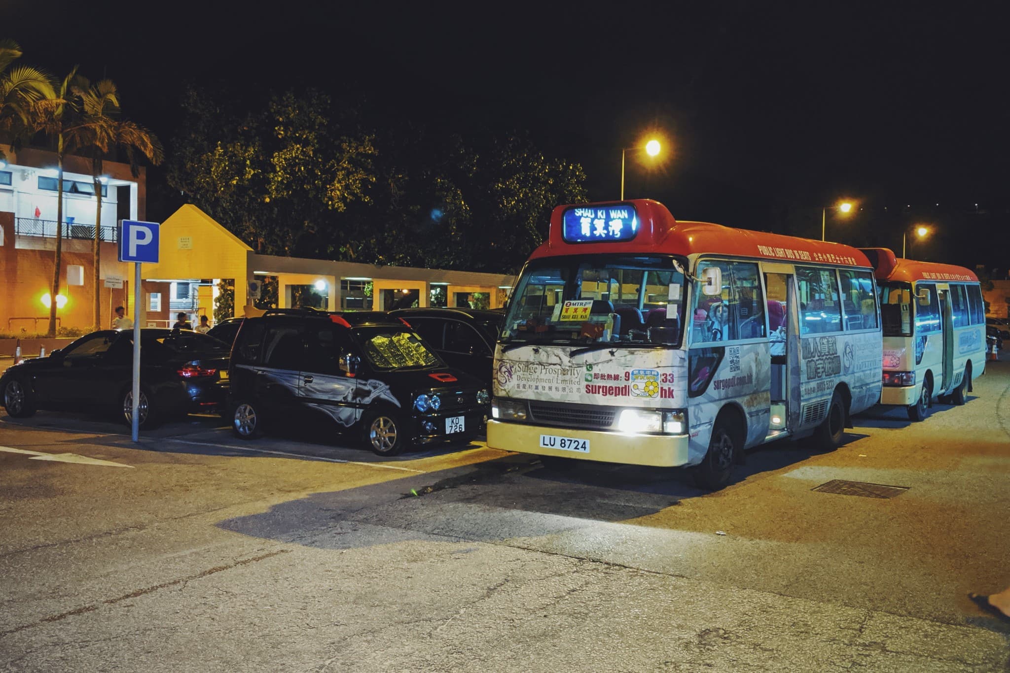 Two red and white buses are parked in a line at night next to several dark cars in a well-lit parking area.