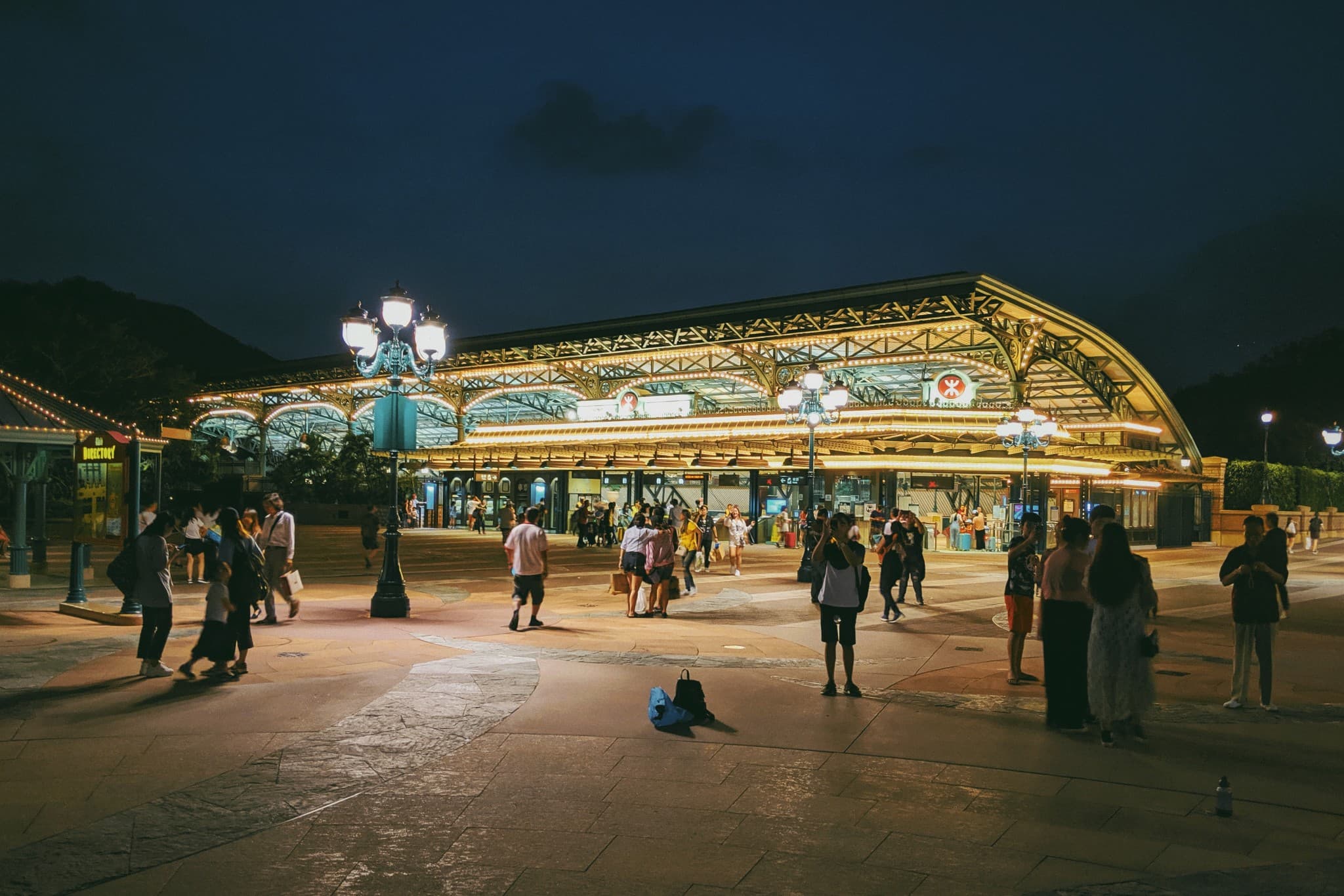 People walk and stand on a paved plaza at night in front of a large, brightly lit station building with an ornate arched roof and vintage streetlamps.