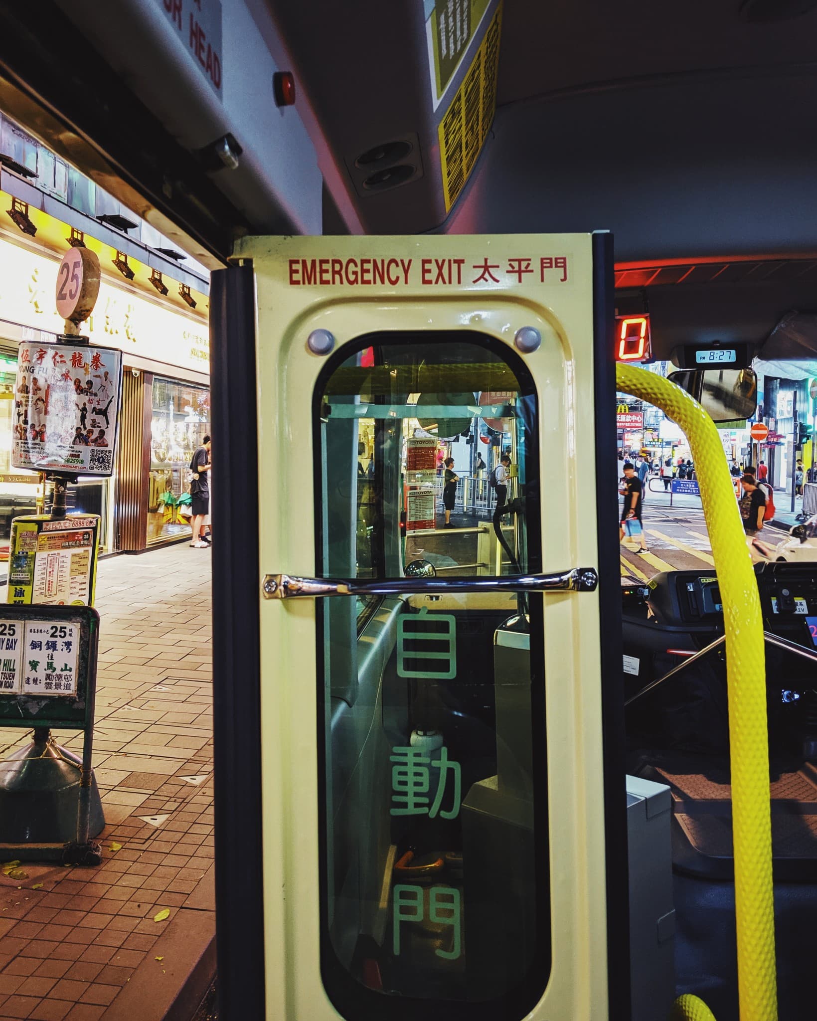 An emergency exit door on a bus, marked with bilingual English and Chinese text, frames a view of a busy city street with shops and pedestrians outside at night.