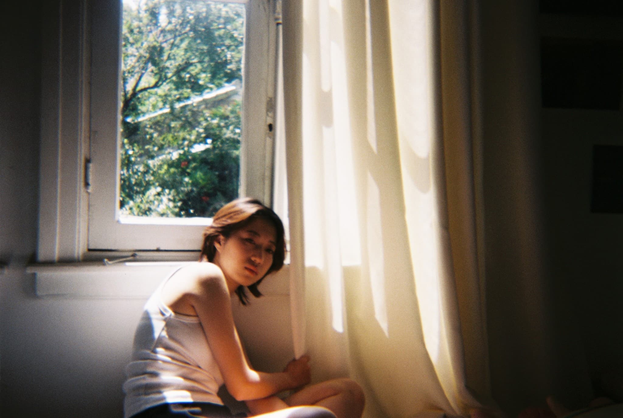 A young woman sits on the floor in a room, illuminated by bright sunlight streaming through a window and sheer curtains.
