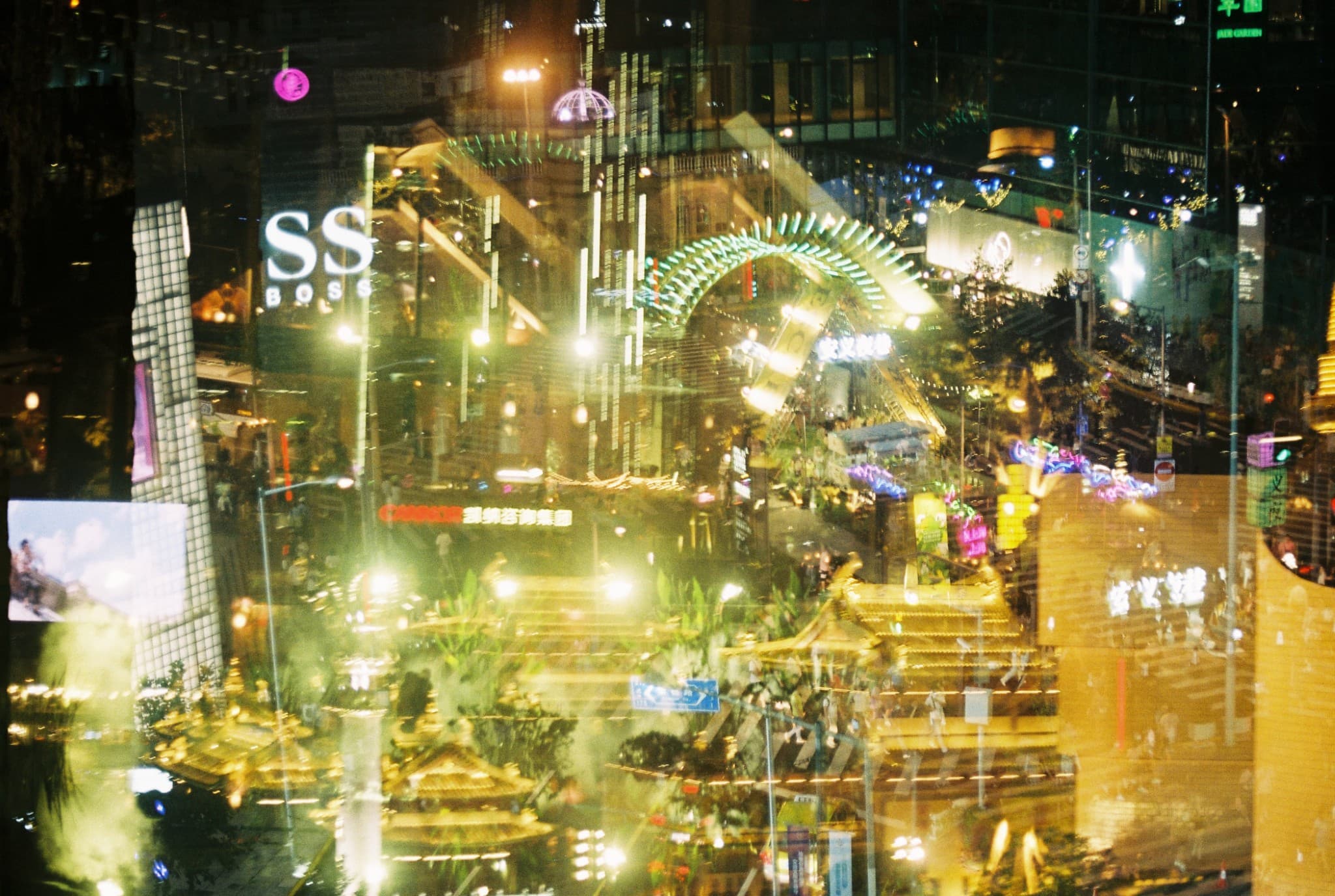 A vibrant, nighttime view of a busy urban shopping district seen through a window, featuring illuminated signs, a green archway, and layered reflections of lights and buildings.