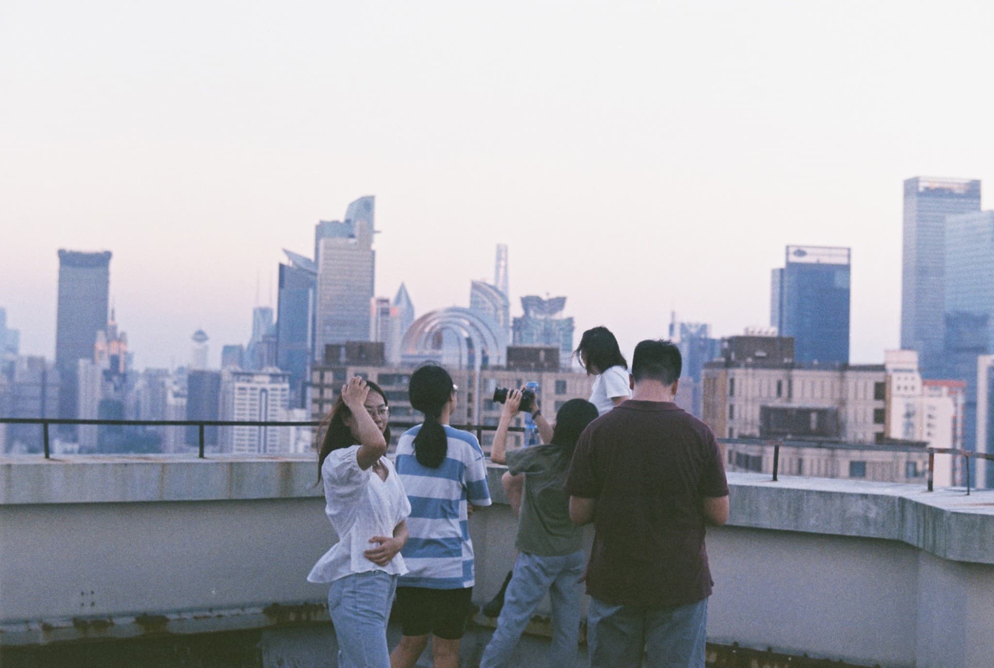A group of people stand on a rooftop terrace overlooking a hazy city skyline filled with tall skyscrapers.