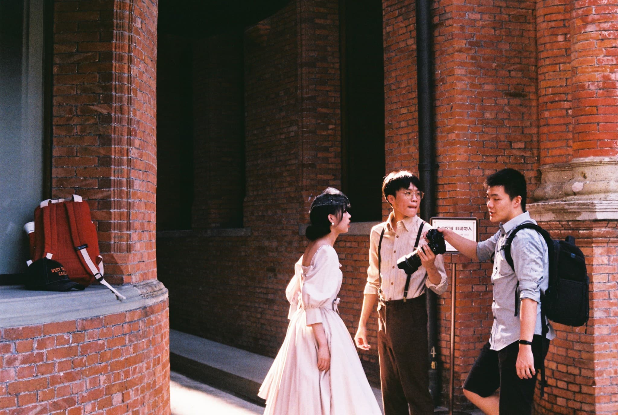 Three young people stand in front of a red brick building, with one man holding a camera while another gestures toward it, and a red backpack rests on a nearby ledge.