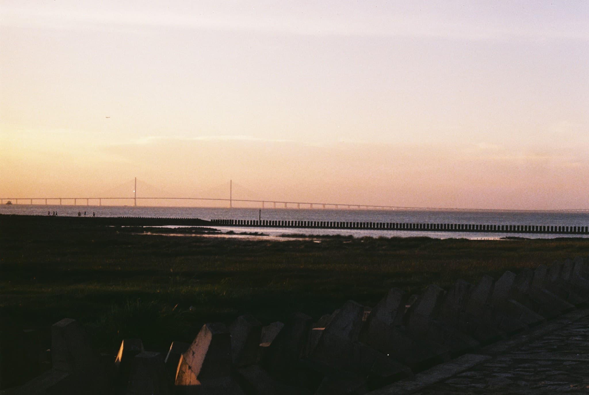A long suspension bridge stretches across the horizon over calm water, viewed from a grassy shore lined with dark concrete blocks under a soft, hazy sunset sky.