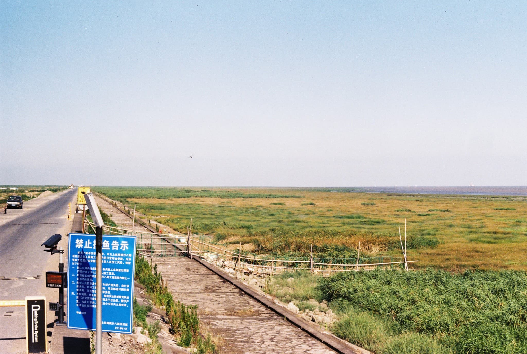 A paved road runs alongside a vast, grassy wetland with a wooden boardwalk and a blue sign featuring Chinese text in the foreground.