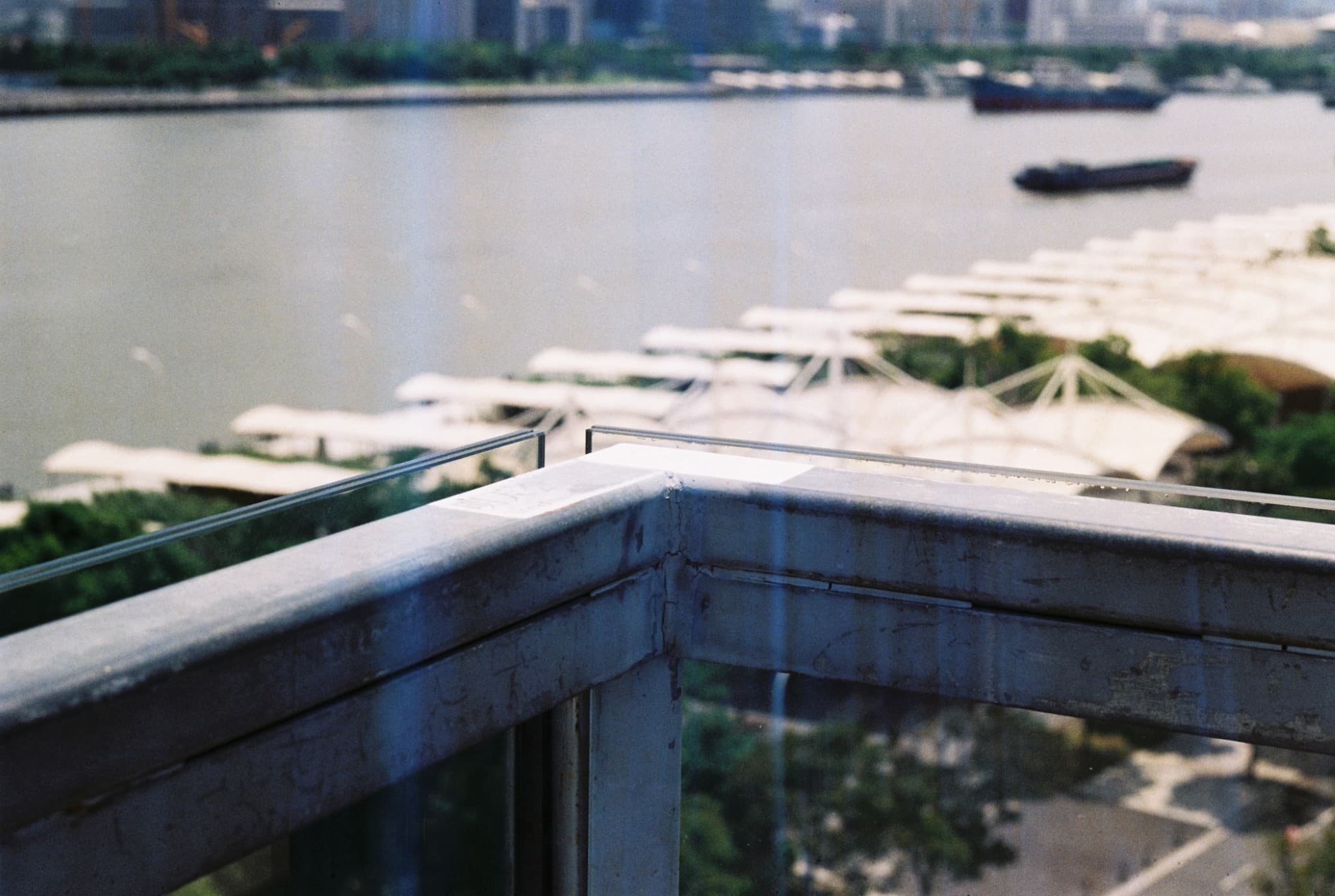 A close-up view of a weathered metal railing overlooking a wide river, where boats and white canopy structures are visible in the distance.