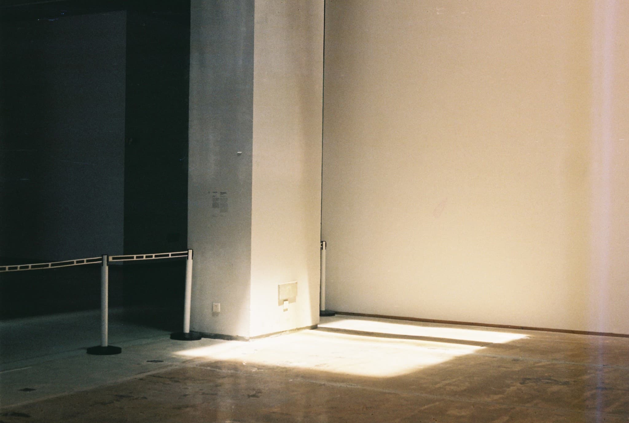 Bright sunlight illuminates the floor and right wall of an empty room, contrasting with a dark, shadowed area on the left near a white column and metal barriers.