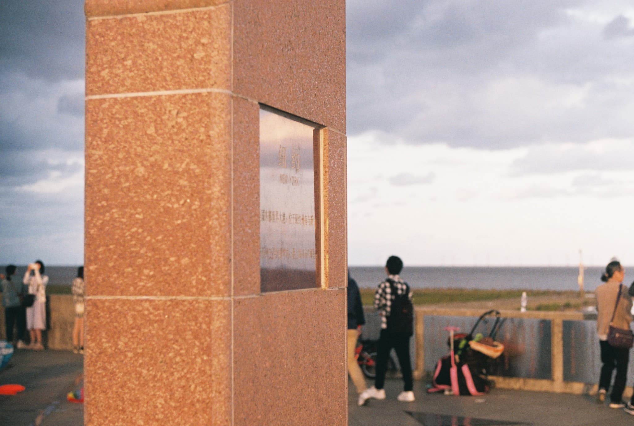 A close-up view of a reddish-brown stone monument with an engraved plaque stands in the foreground, set against a blurred background of a cloudy sky, the sea, and people standing near a railing.