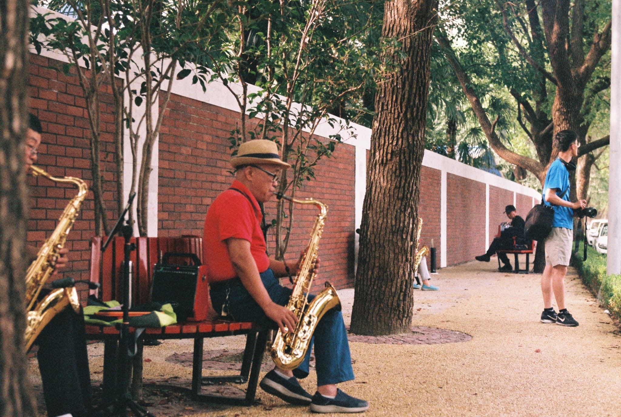 A man in a red shirt and hat plays the saxophone on a park bench next to another musician, while a photographer stands nearby on a gravel path.
