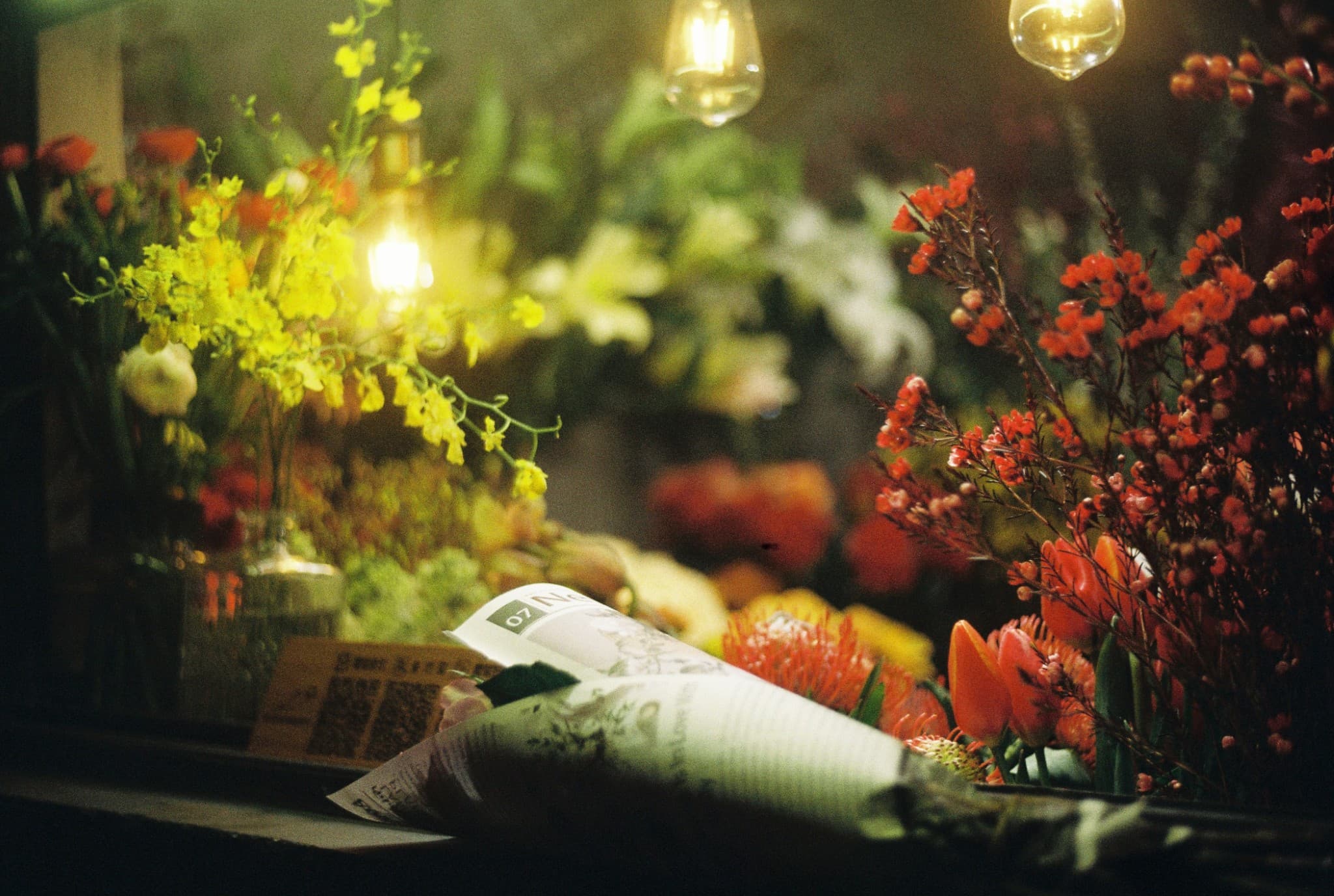 A wrapped bouquet rests in the foreground of a flower display featuring yellow orchids on the left and red flowers on the right, illuminated by warm hanging light bulbs.