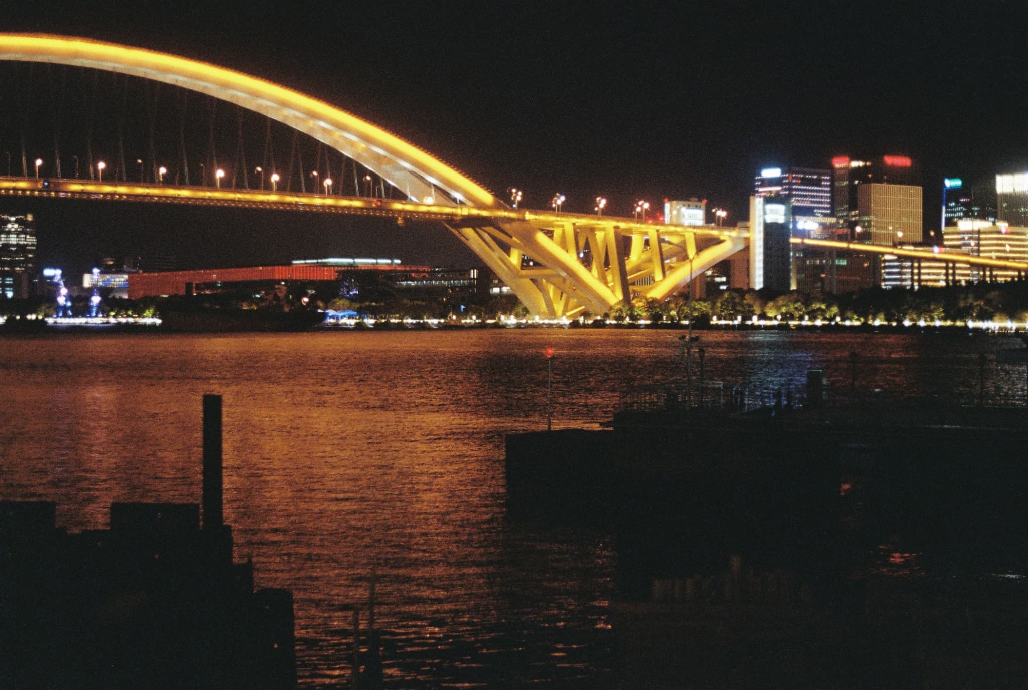 A large, brightly lit yellow bridge spans a river at night, with its lights reflecting on the water and a city skyline visible in the background.