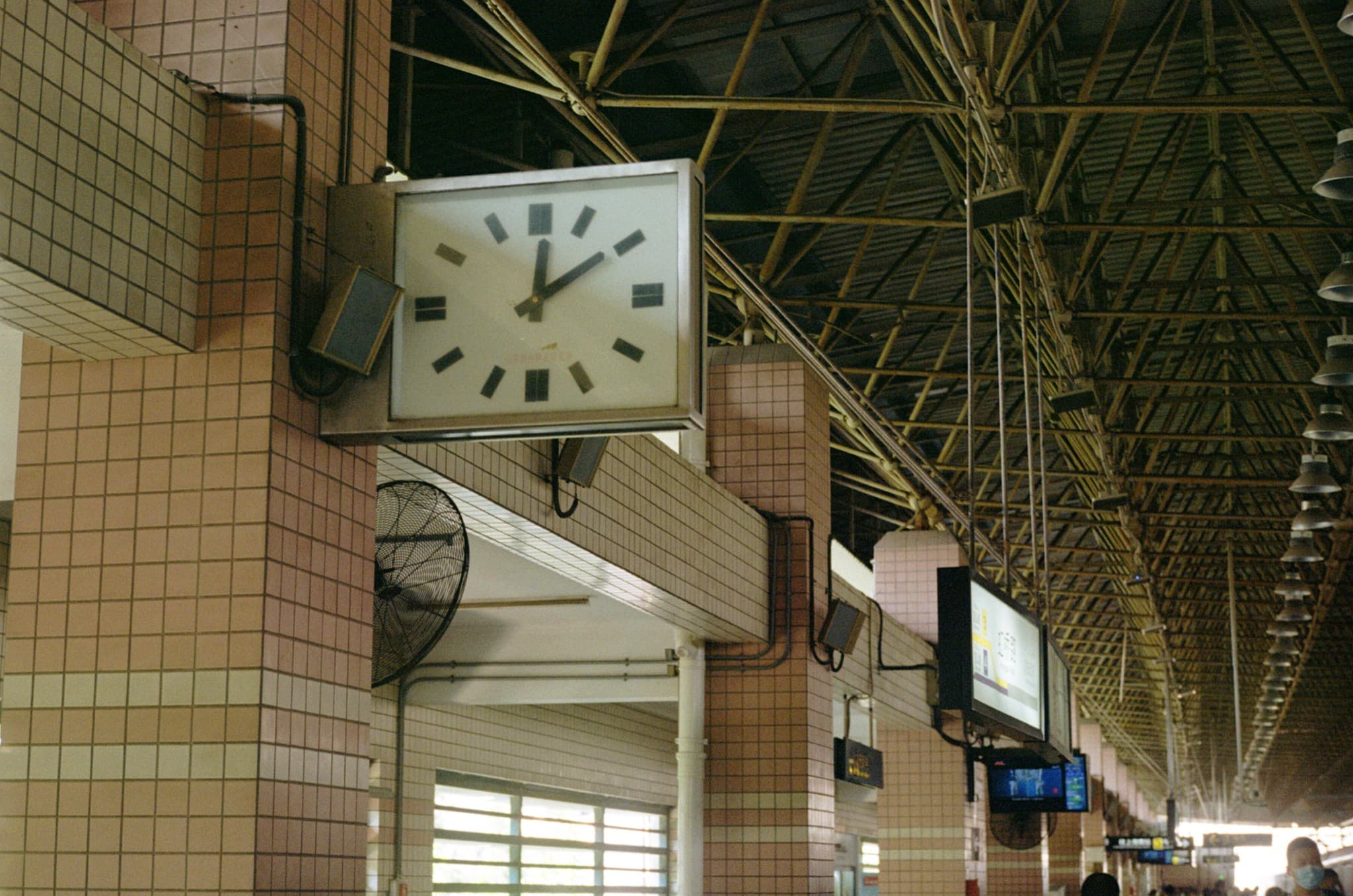 A large square analog clock hangs from the high, truss-supported ceiling of a tiled train station platform.