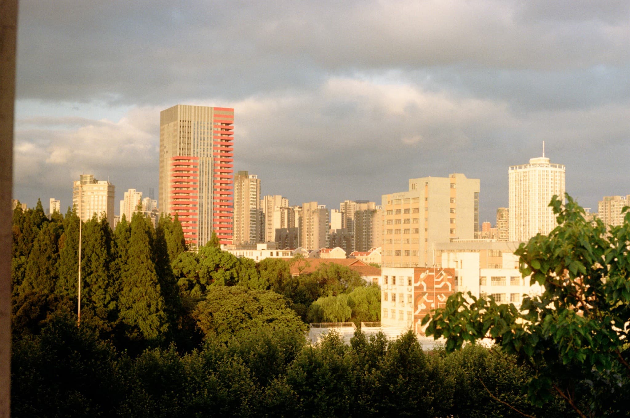 Green trees in the foreground frame a view of a city skyline featuring a prominent red and grey skyscraper under a cloudy sky.