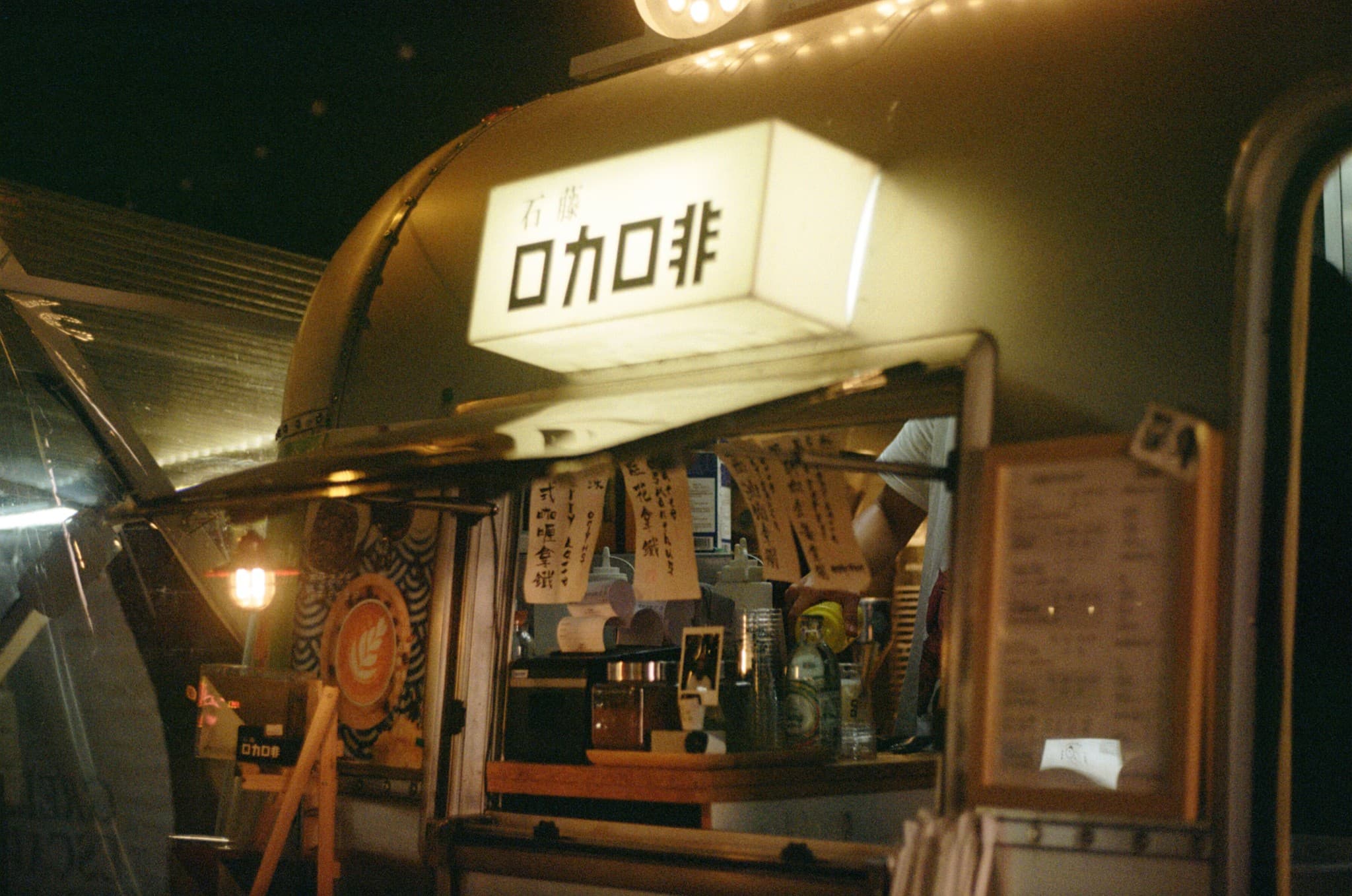 An illuminated mobile coffee cart at night features a large sign with Japanese characters and an open service window revealing bottles and cups inside.