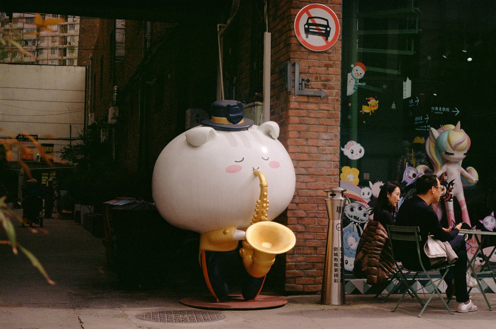 A large white statue of a character wearing a top hat plays a saxophone on the sidewalk next to a brick wall and a window decorated with colorful cartoon figures.