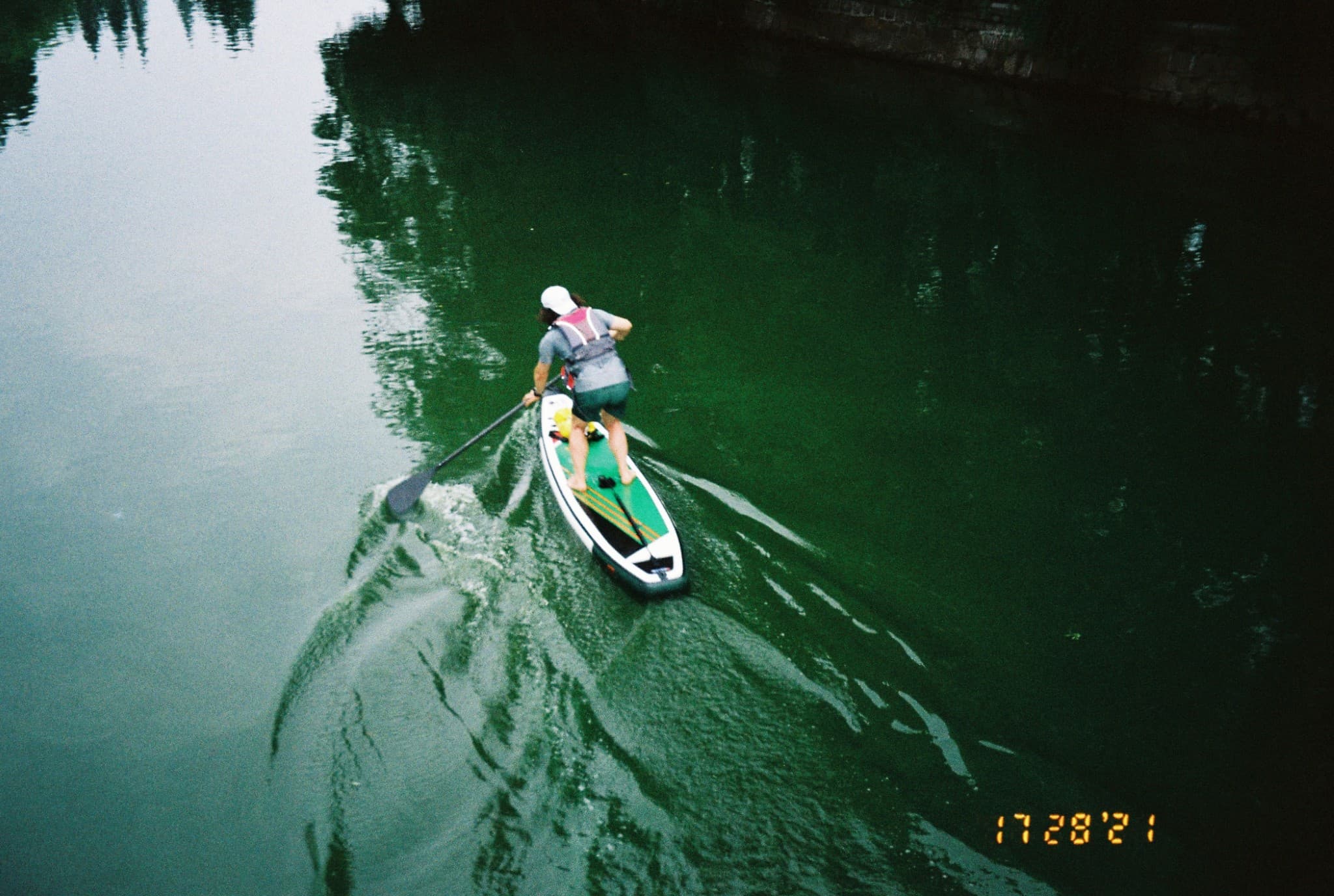 A person wearing a life vest and white hat paddleboards down a green river, leaving a wake in the water.