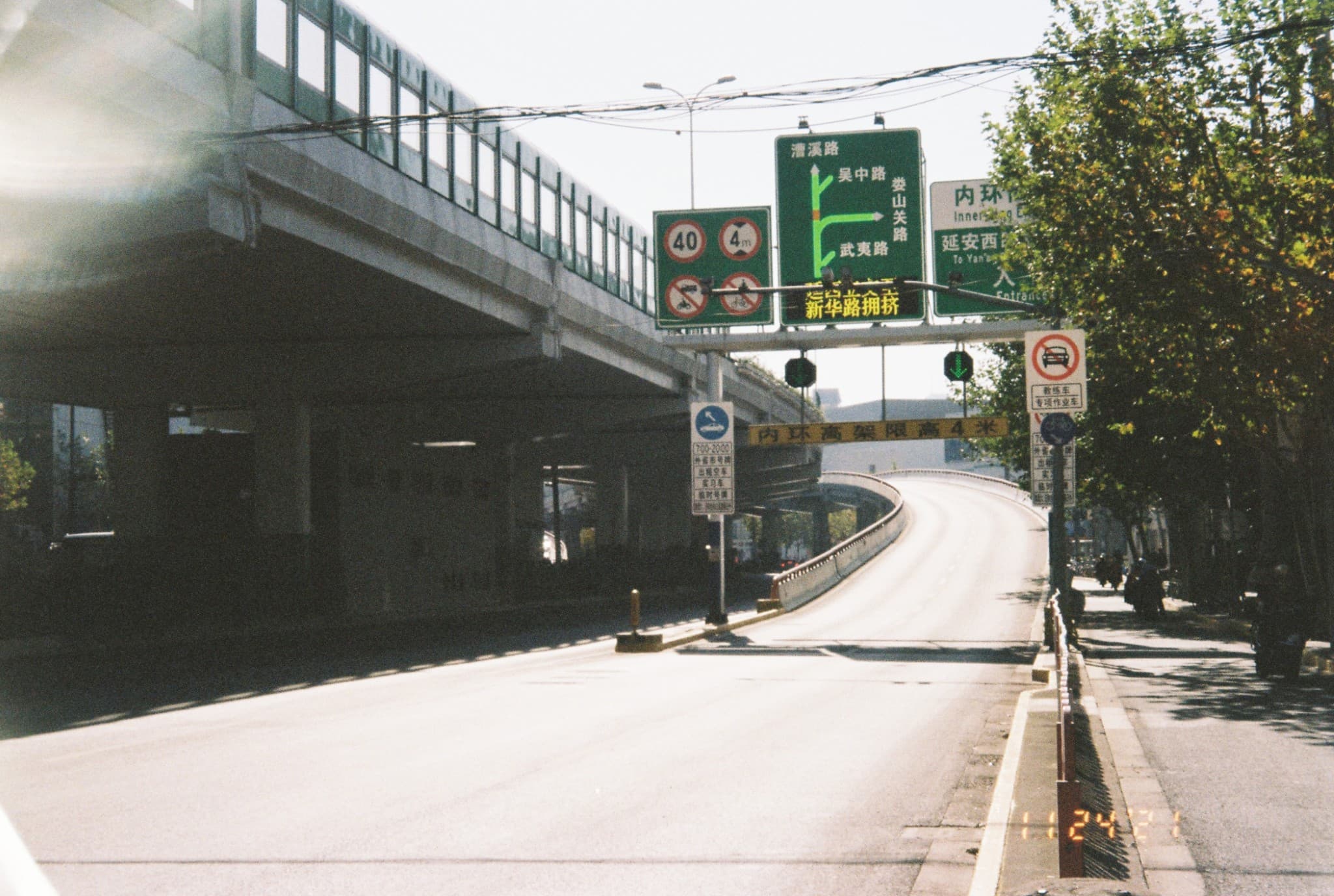 A view of an empty road ramp curving upward beneath a concrete overpass, marked by overhead green directional signs and restriction placards.