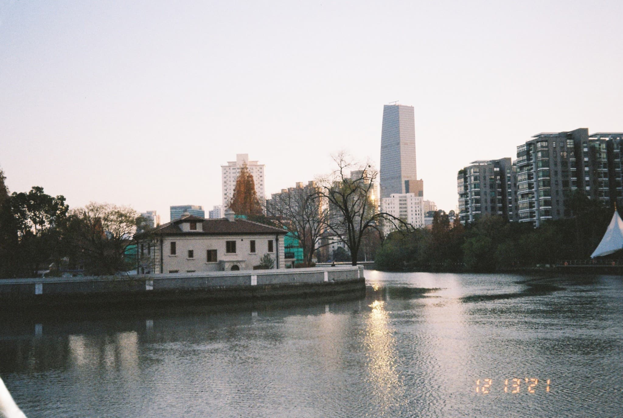 A calm river reflects the sunlight, bordered by a low white building and trees on the left bank, with a backdrop of modern city skyscrapers.