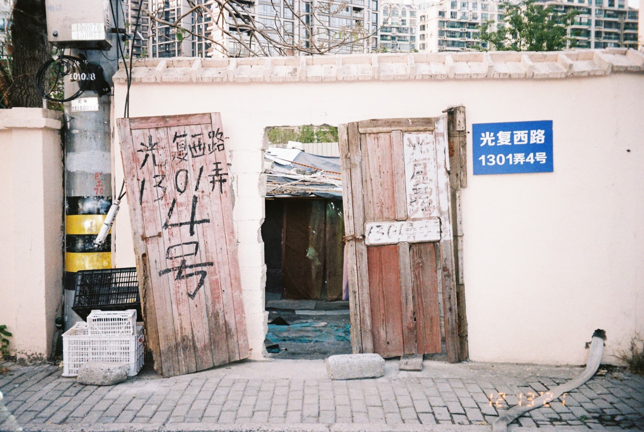 A weathered wooden door with handwritten Chinese characters leans against a cream-colored wall next to a blue street sign marking an alleyway entrance.