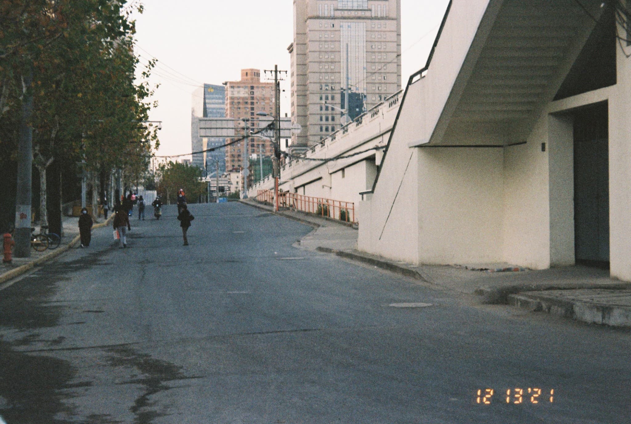 A paved road stretches uphill between a sidewalk with walkers and trees on the left, and a white building with an overhang on the right, set against a backdrop of high-rise buildings.