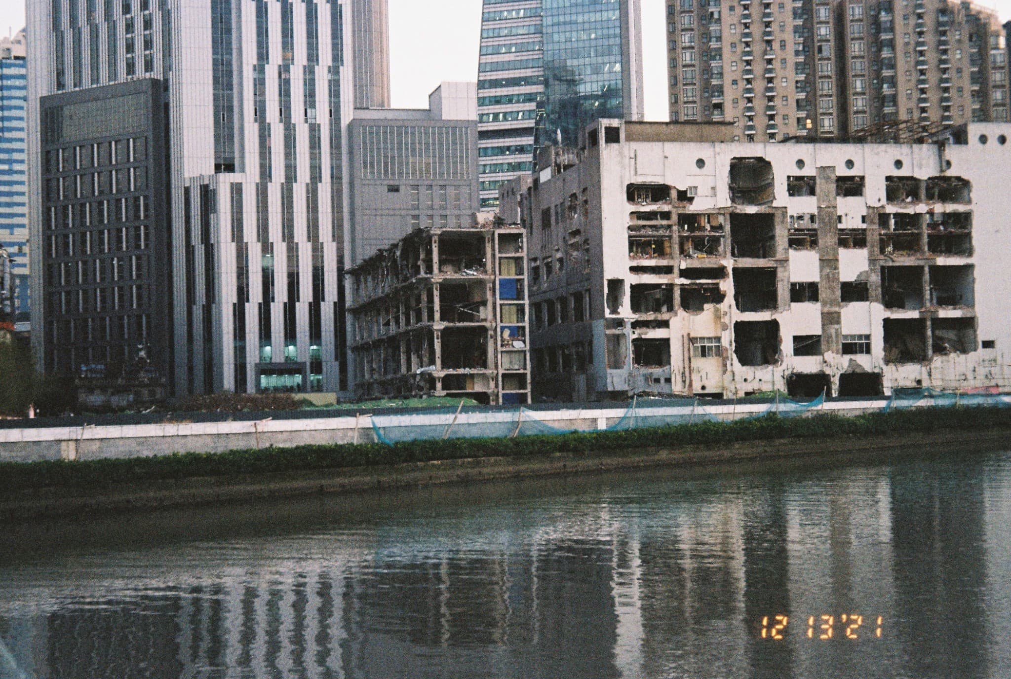 A row of dilapidated, partially demolished buildings stands along a riverbank, contrasting with the tall, modern skyscrapers in the background.
