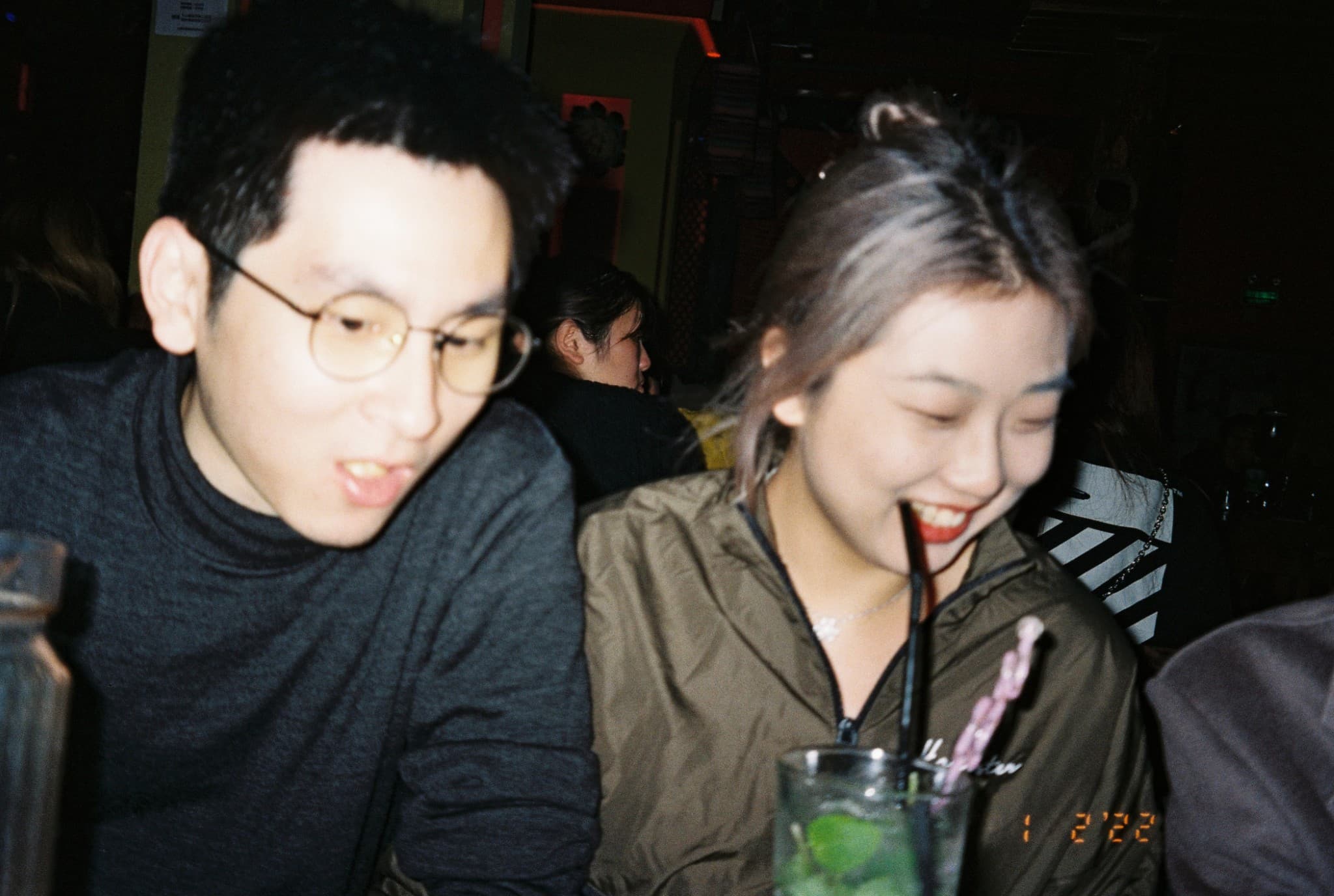 A young man with glasses and a dark sweater sits next to a smiling young woman in a green jacket, both looking down in a dimly lit setting with a cocktail glass in the foreground.