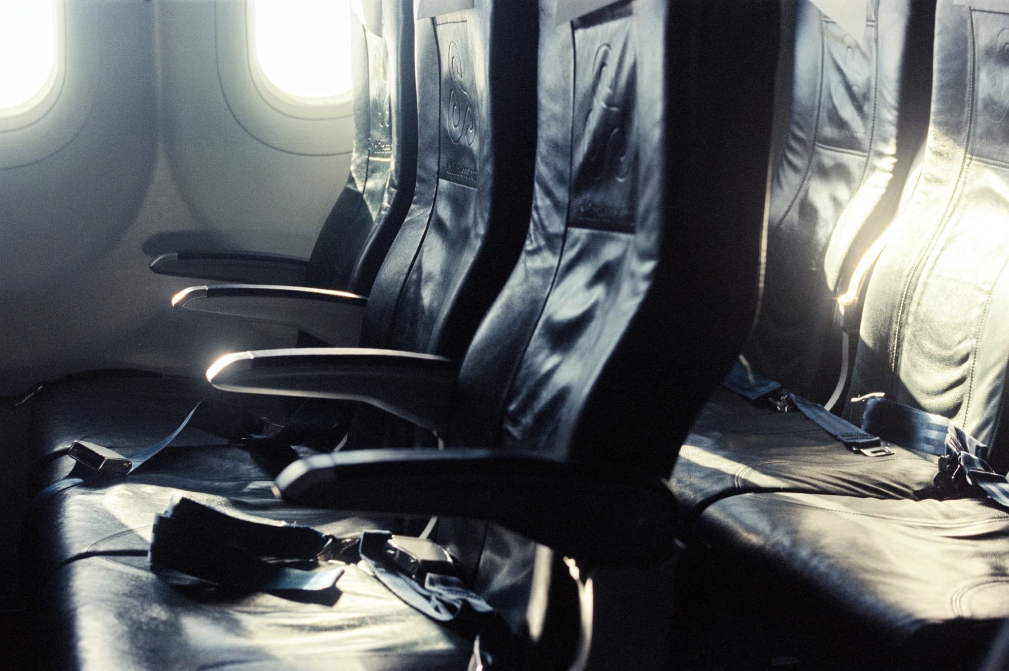 Rows of empty, dark airplane seats with loose safety belts are illuminated by bright sunlight streaming through the windows.