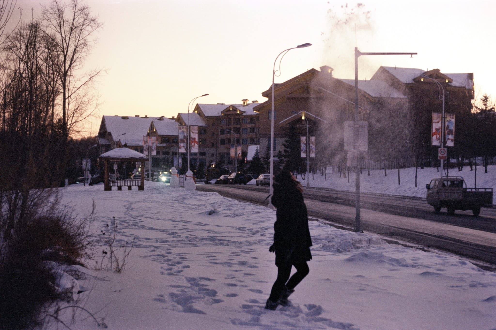 A person walks along a snow-covered path near a road with large buildings and streetlights in the background during the winter twilight.