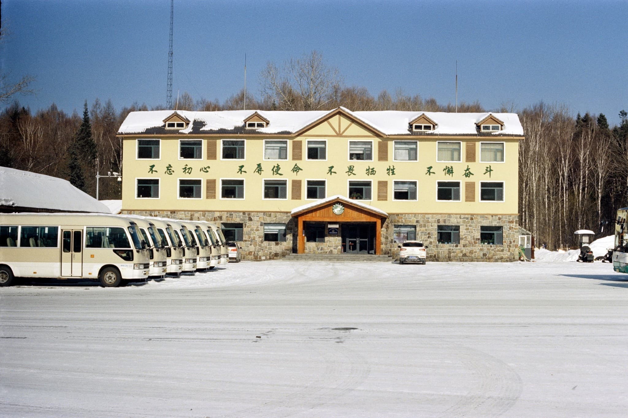 A yellow building with Chinese text on the facade stands in a snowy lot next to a row of parked white buses.