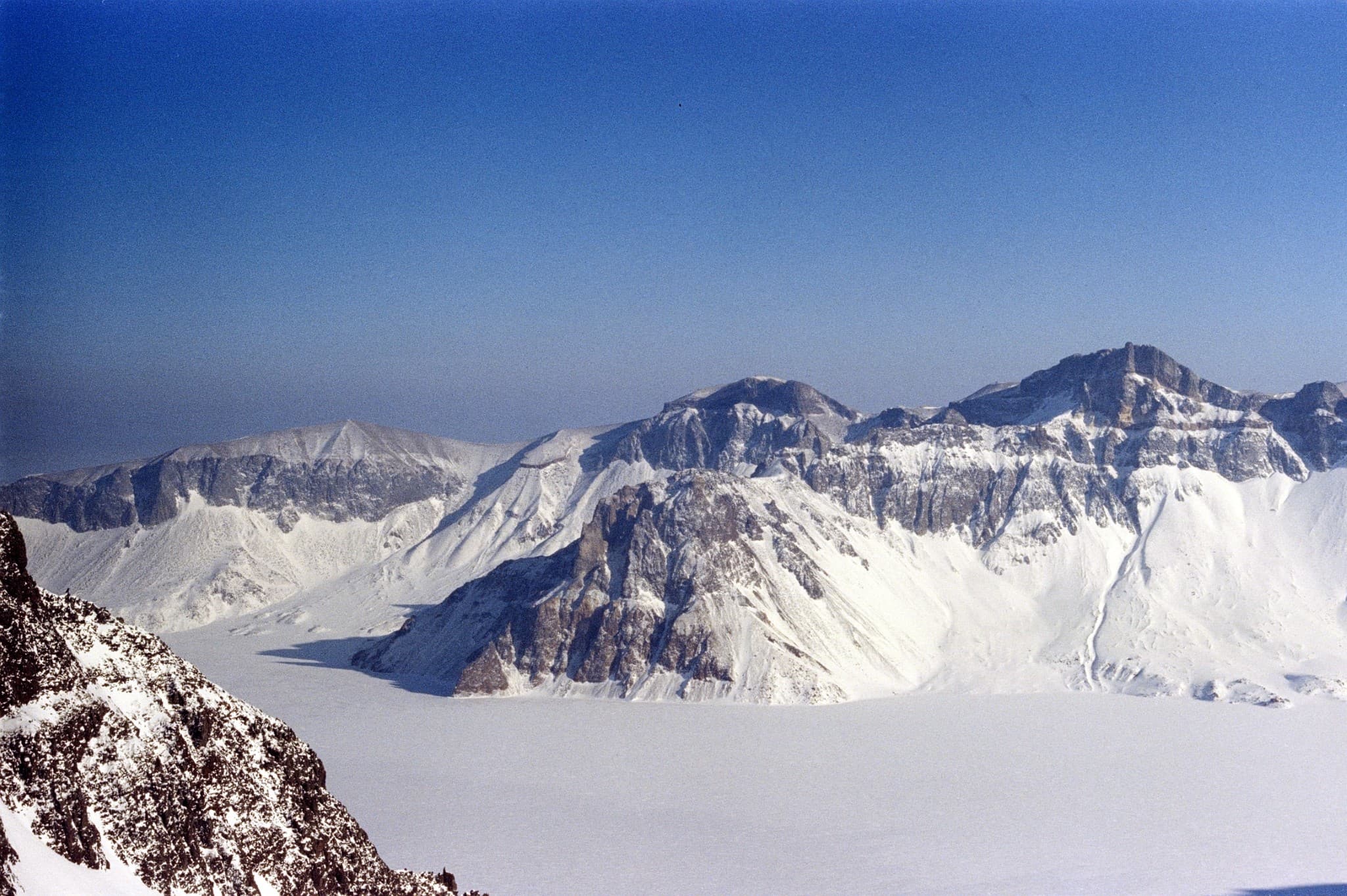 Rugged, snow-covered mountain peaks and rocky cliffs overlook a vast, flat white landscape under a clear blue sky.