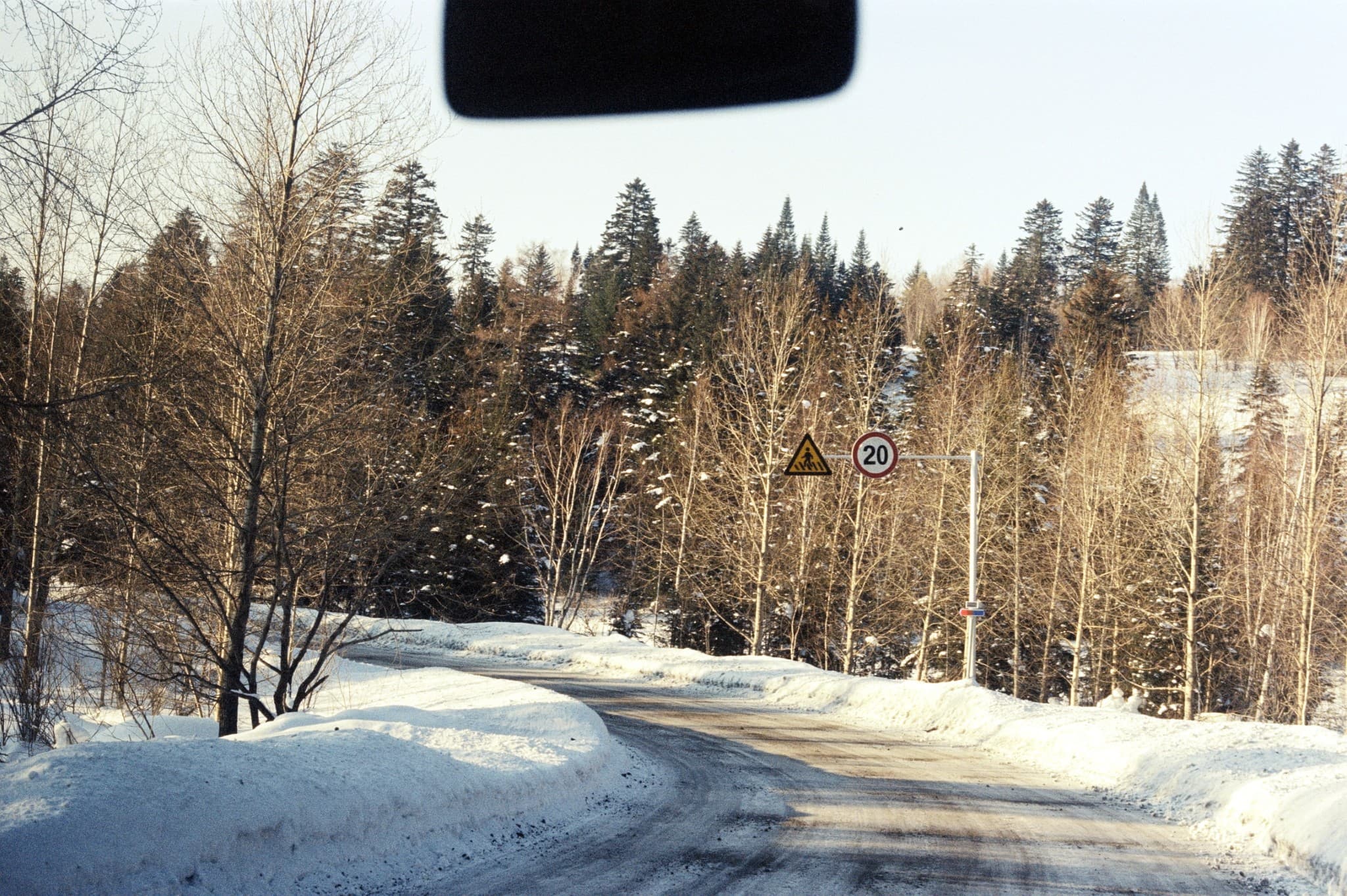 A view from inside a vehicle looking out onto a snow-covered road curving through a winter forest, marked by a sign indicating a speed limit of 20.