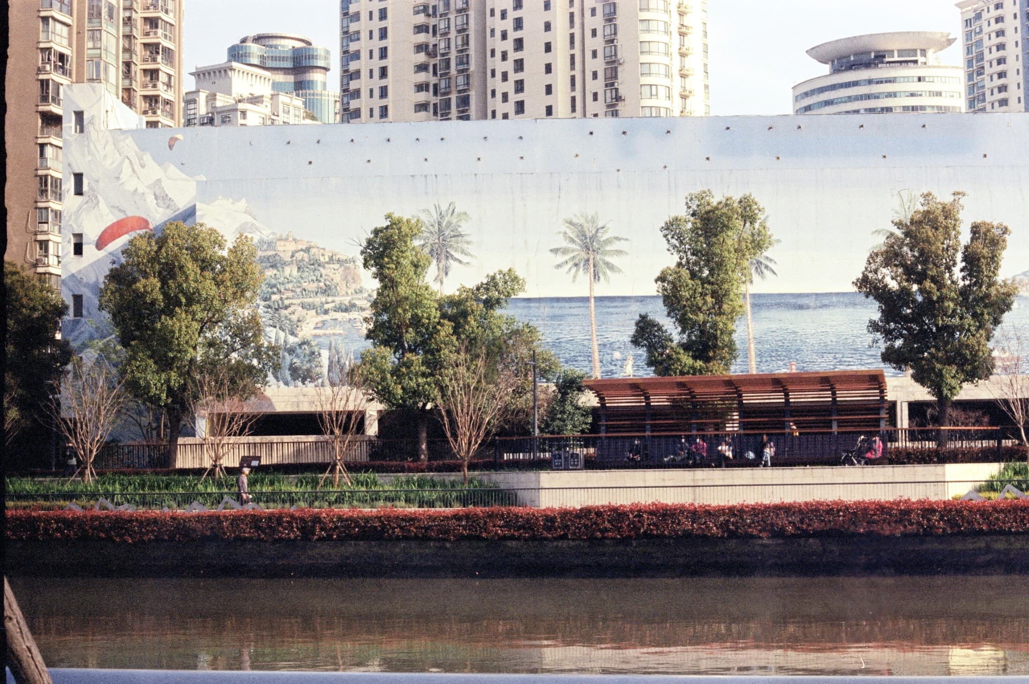 A large mural depicting a tropical beach scene with palm trees and mountains is painted on a wall in an urban setting, partially obscured by real trees and bushes.